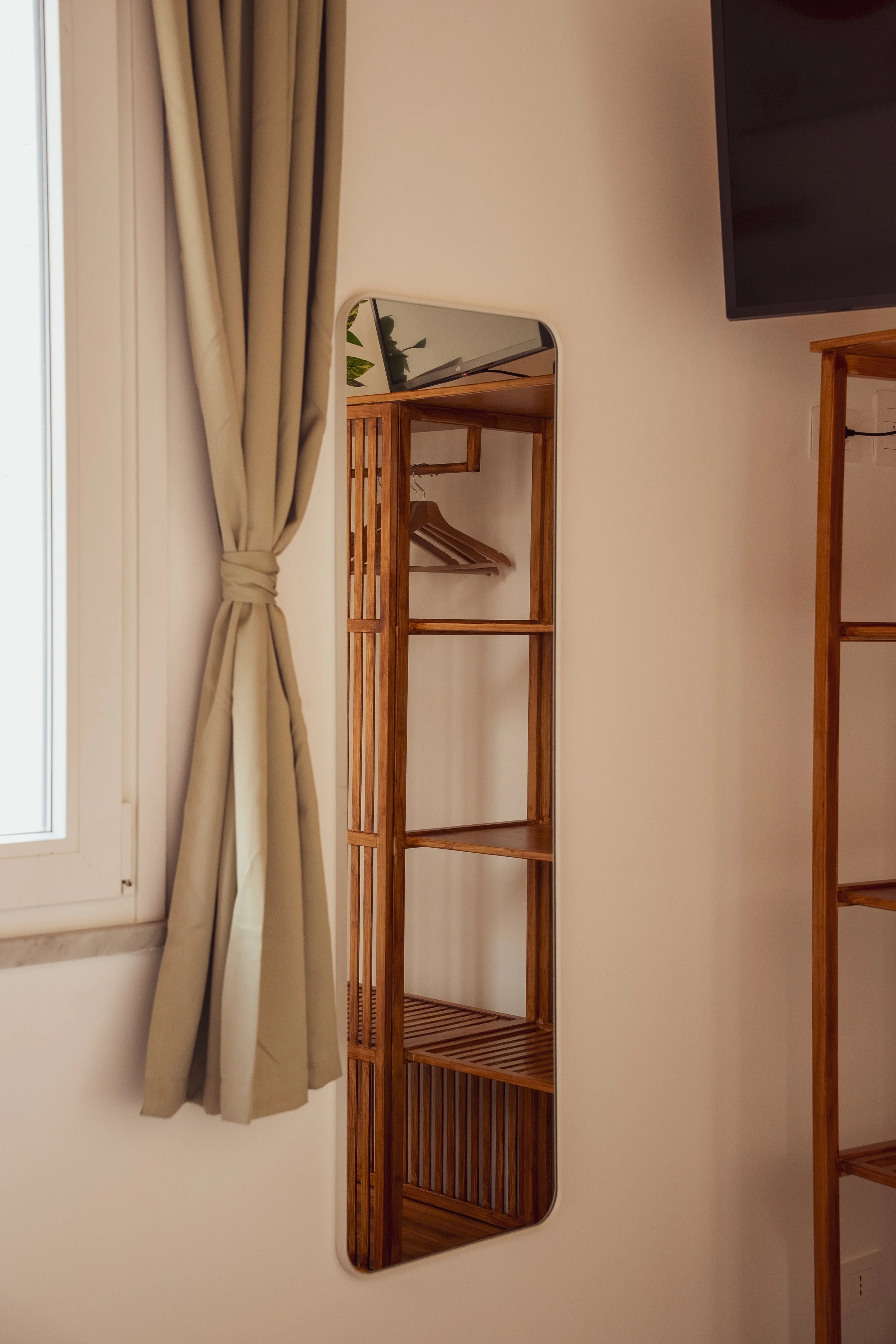 Specchio a parete riflettente accanto a una finestra con tenda beige e scaffale in legno