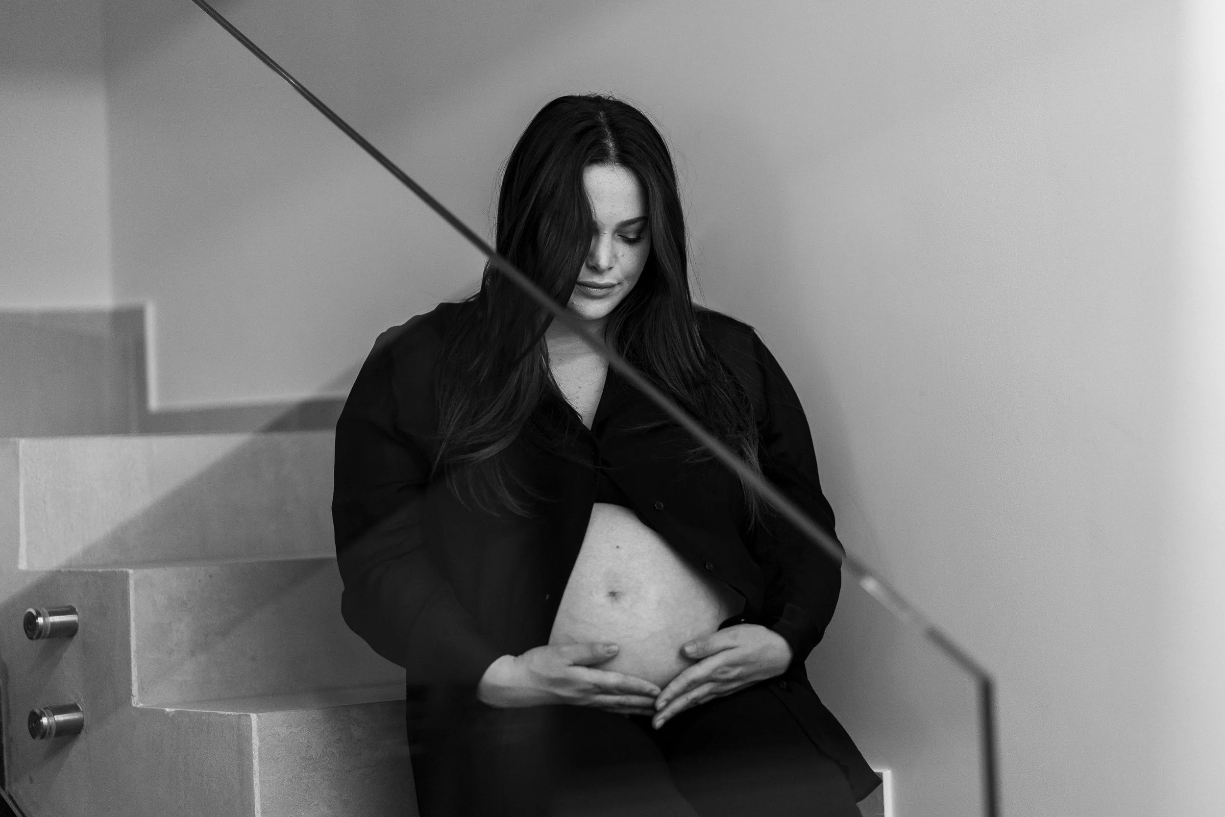 Ragazza incinta seduta su le scale, con le mani sul ventre, vestita di nero, con i capelli castani lunghi, in bianco e nero.