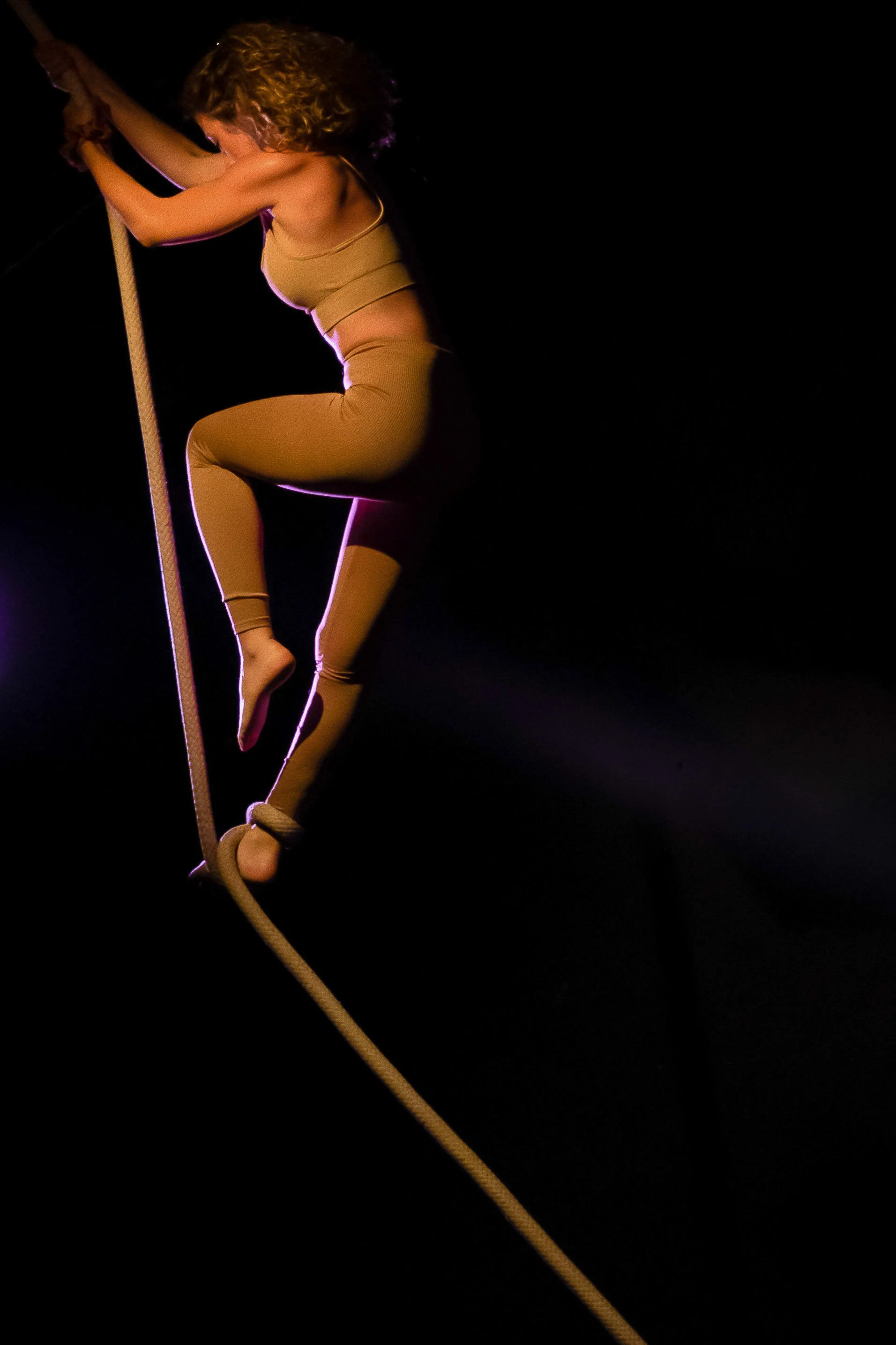 Performing artist in beige outfit balancing on a tightrope with a dark background.