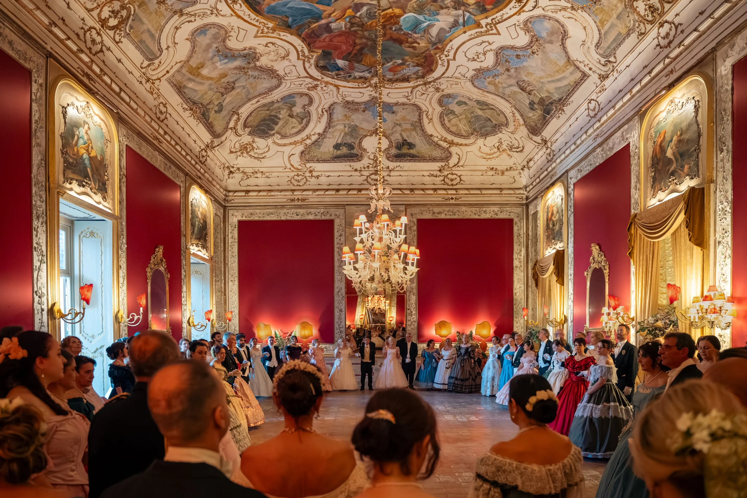 Salone elegante con soffitto affrescato e lampadario chandelier, molte persone vestite in abiti storico-romantici, probabilmente durante un ballo o matrimonio.