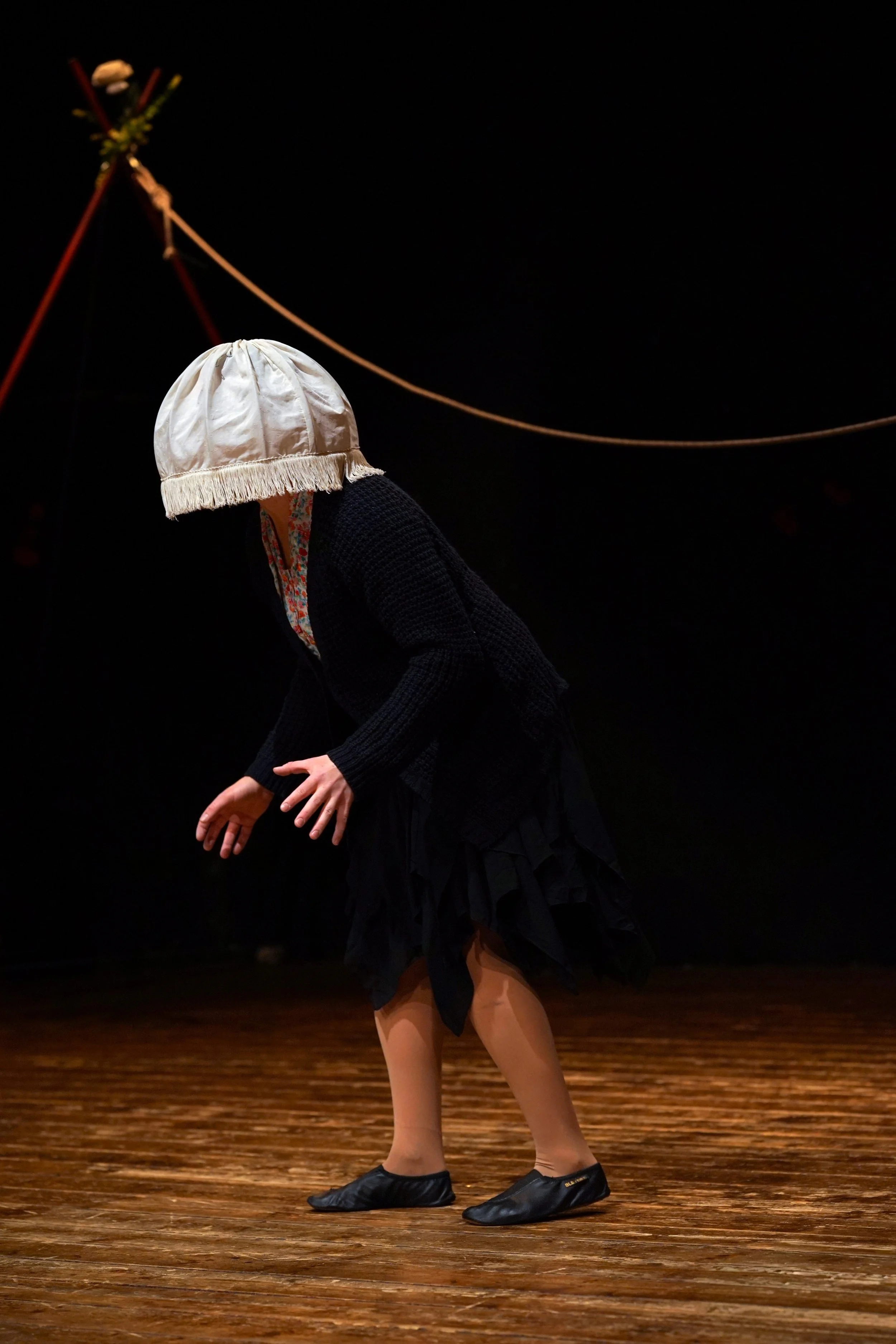 Una persona con un cappello di tessuto bianco, vestendo un abbigliamento nero, si esibisce su un palco di legno scuro, con uno sfondo nero e una struttura rossa decorativa sullo sfondo.