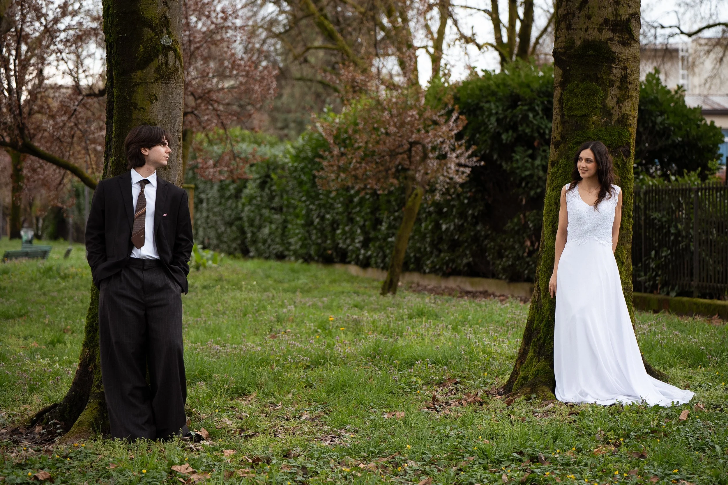 Un uomo e una donna in abiti eleganti si trovano in un parco con alberi che hanno fiori primaverili, il uomo ha i capelli castani corti e indossa un completo nero con una cravatta, la donna ha capelli scuri ondulati e indossa un abito bianco lungo da