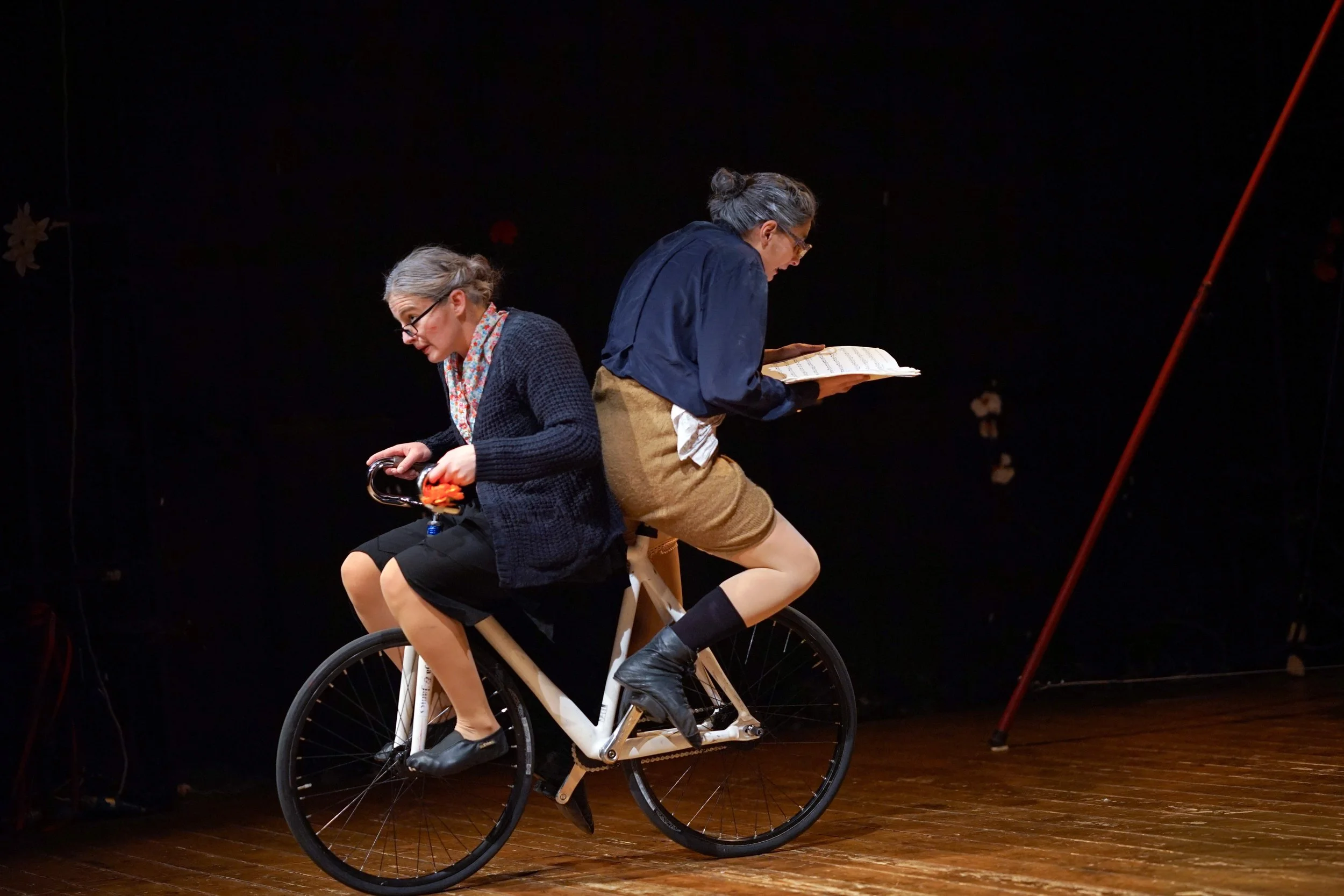 Due donne anziane, una seduta e una in piedi, che si trovano su una bicicletta in un ambiente con pavimento in legno e pareti nere. La donna seduta tiene un cappello, mentre la donna in piedi leggendo un libro si trova dietro di lei.