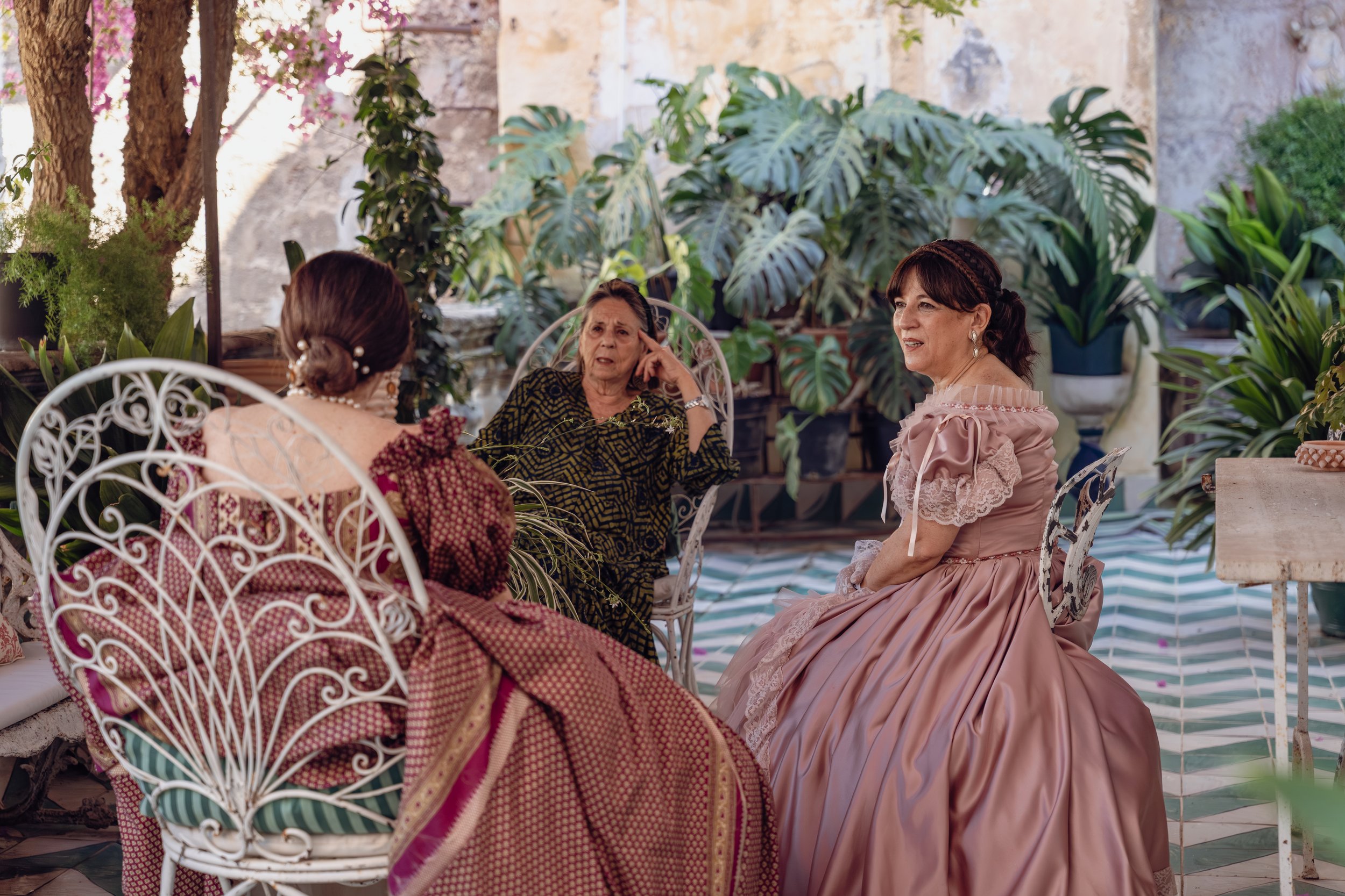 Quattro donne vestite con abiti eleganti si siedono in un giardino lussureggiante con piante verdi e grandi foglie, mentre conversano.