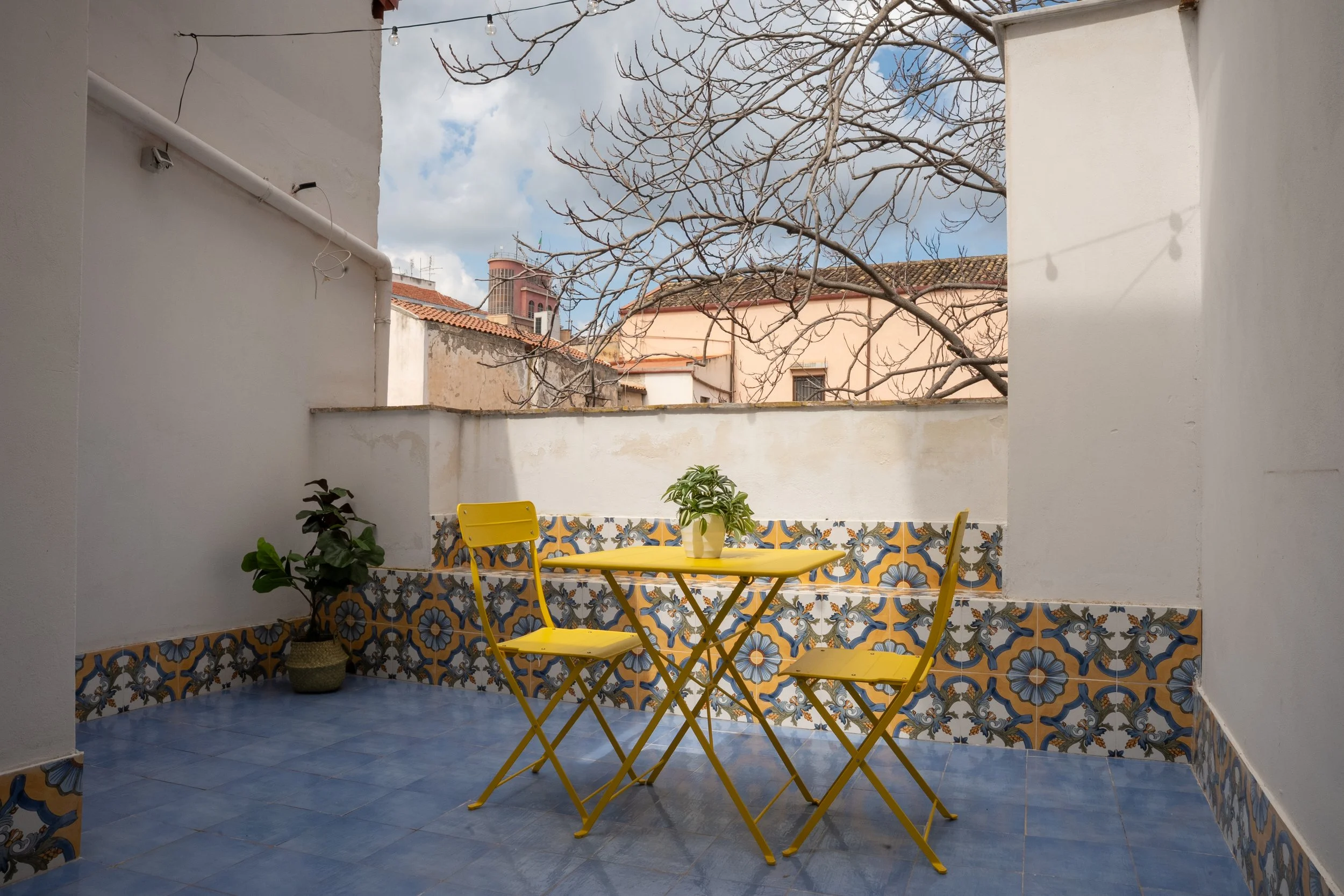 Terrazza con pianta, tavolo e due sedie gialle, decorazioni di piastrelle colorate, cielo con nuvole, edifici e alberi con rami senza foglie sullo sfondo.