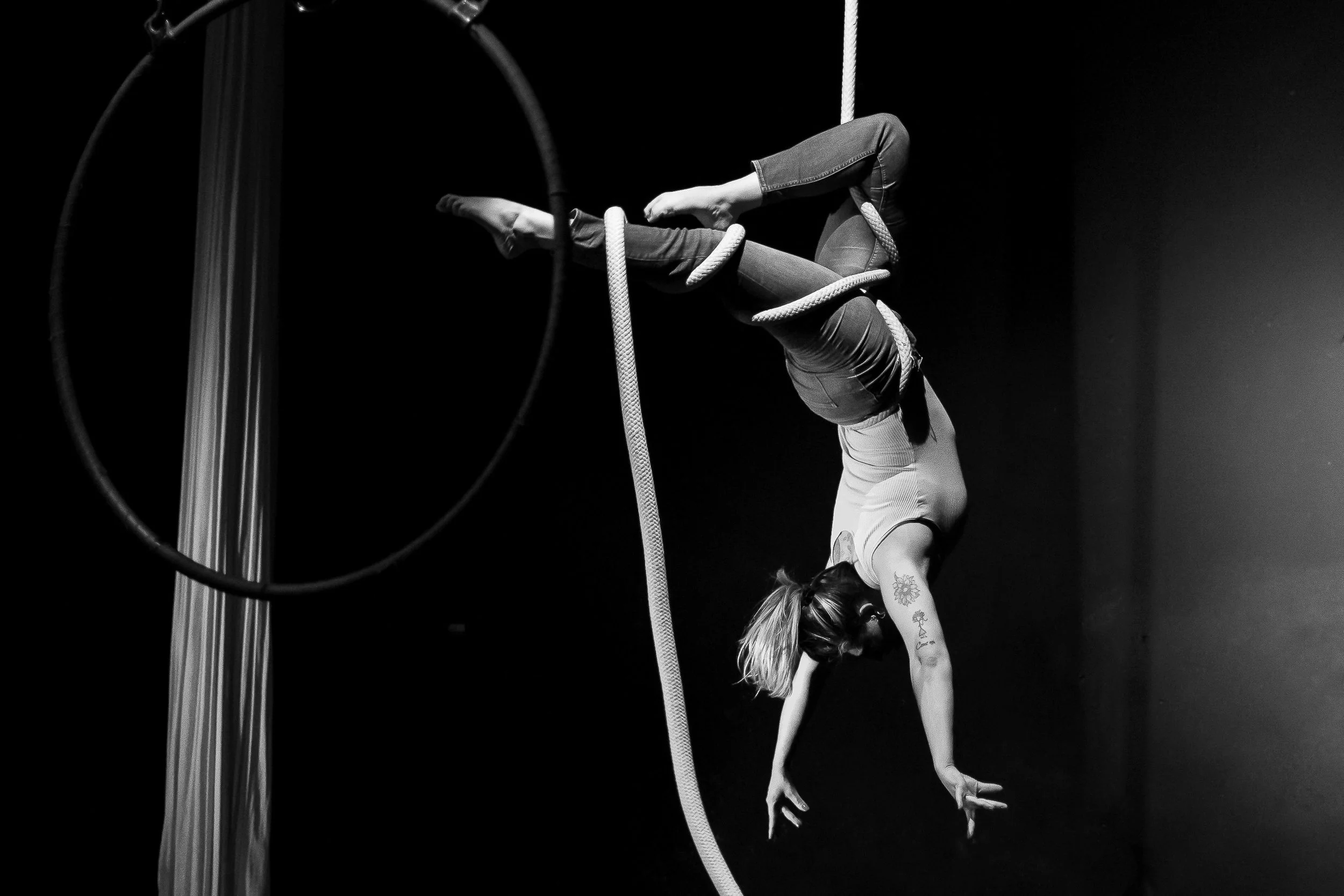 Una donna esegue una performance acrobatica in aria usando una corda e un cerchio. È capovolta con le mani a terra e le gambe appoggiate sulla corda, mentre si tiene in equilibrio.