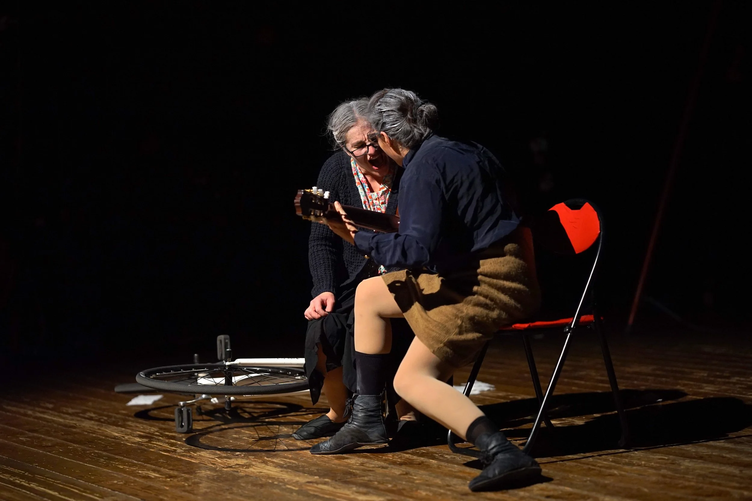 Due attori in scena, uno con in mano un libro, l'altro seduto su una sedia rossa, su un palco di legno, in un'illuminazione concentrata.