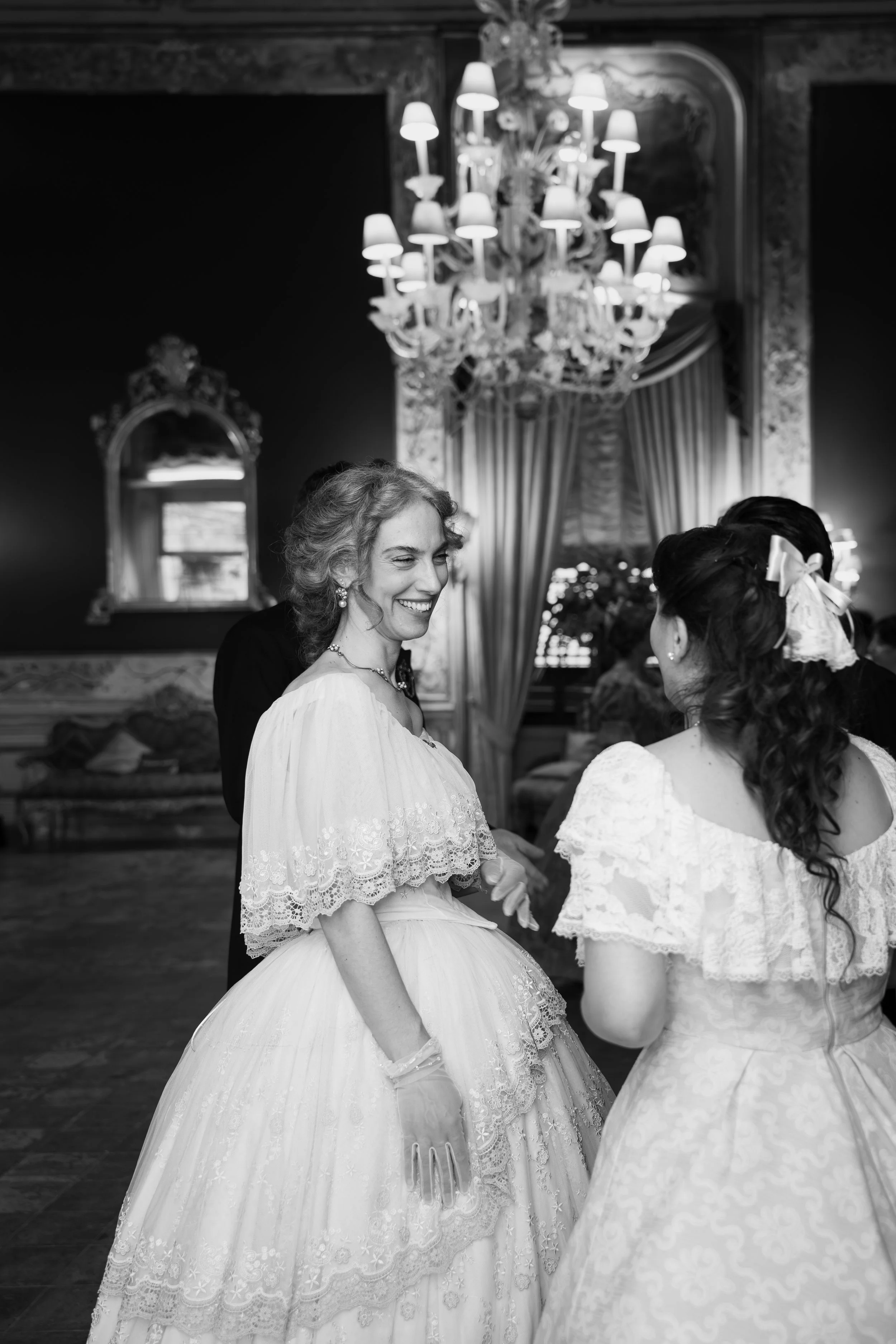 Due donne in abiti da cerimonia sorridenti mentre si scambiano promesse o promesse di matrimonio, in un ambiente elegante con un grande lampadario e decorazioni ornate alle pareti.