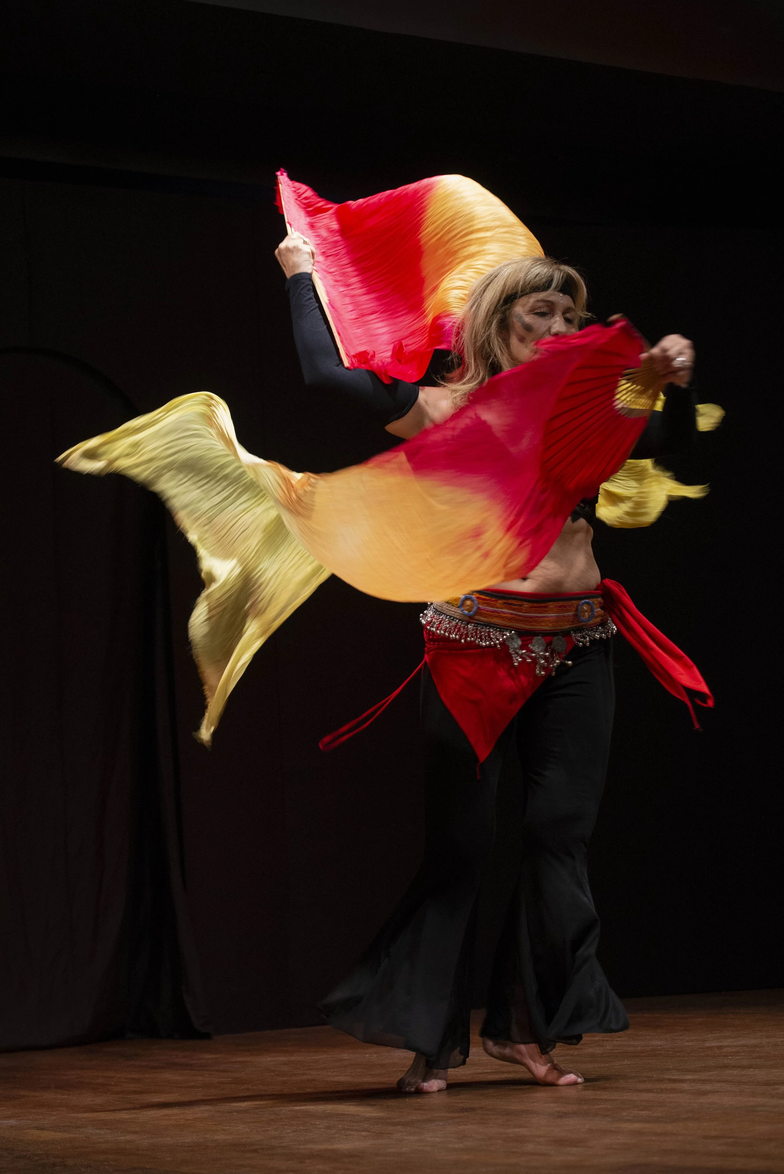 Una donna che danza con scialli colorati mentre si esibisce su un palco di legno, con uno sfondo nero.