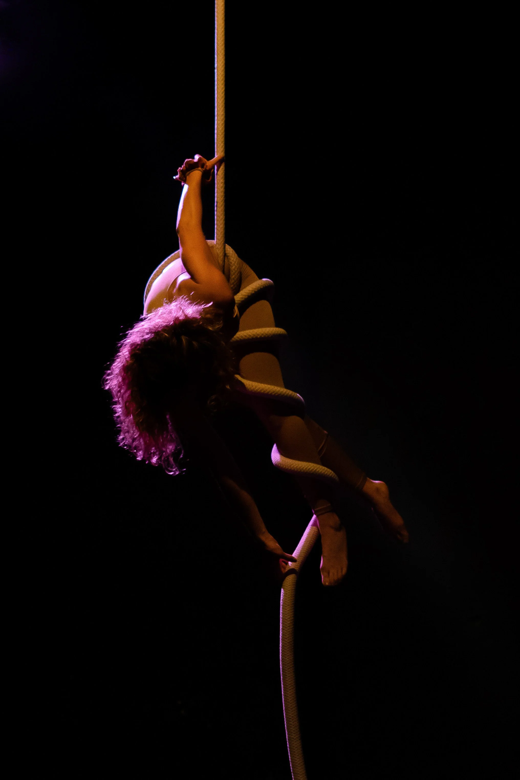 Una donna si esibisce in una performance acrobatica appesa a una corda su un palco oscuro, con luci soffuse