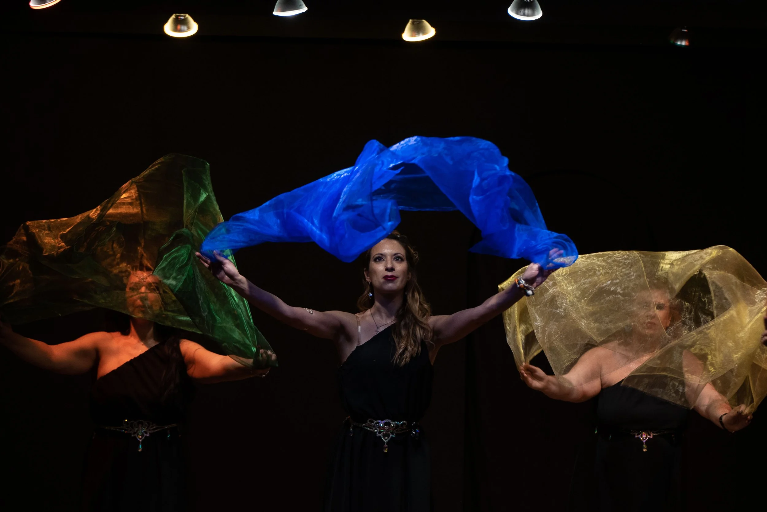 Tre donne in abiti neri che ballano con veli colorati (giallo, verde e blu) su un palco con sfondo nero e luci sopra.