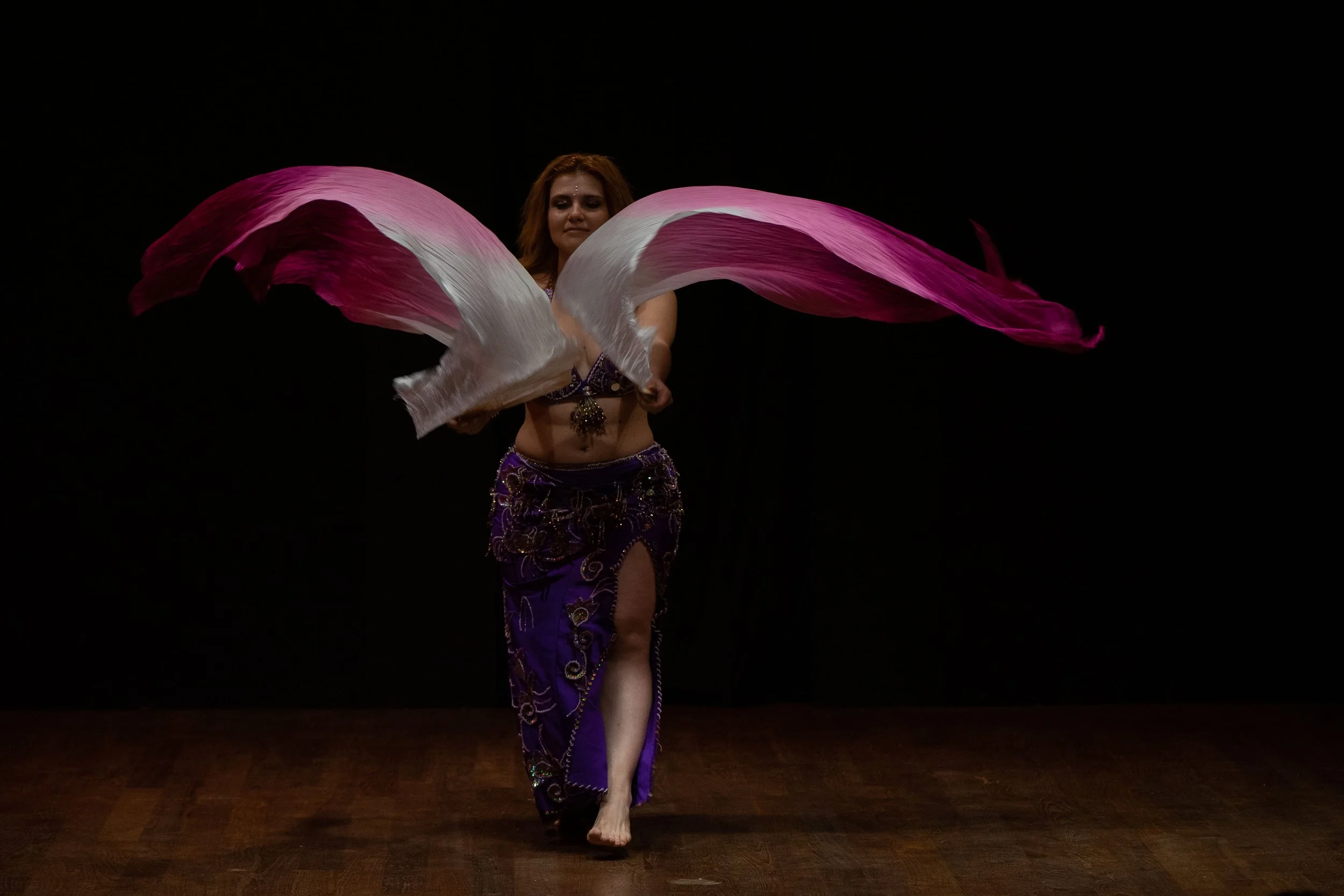 una ballerina che esegue una danza con tessuti rosa e bianchi, indossa un costume viola decorato, sul palco con sfondo nero.