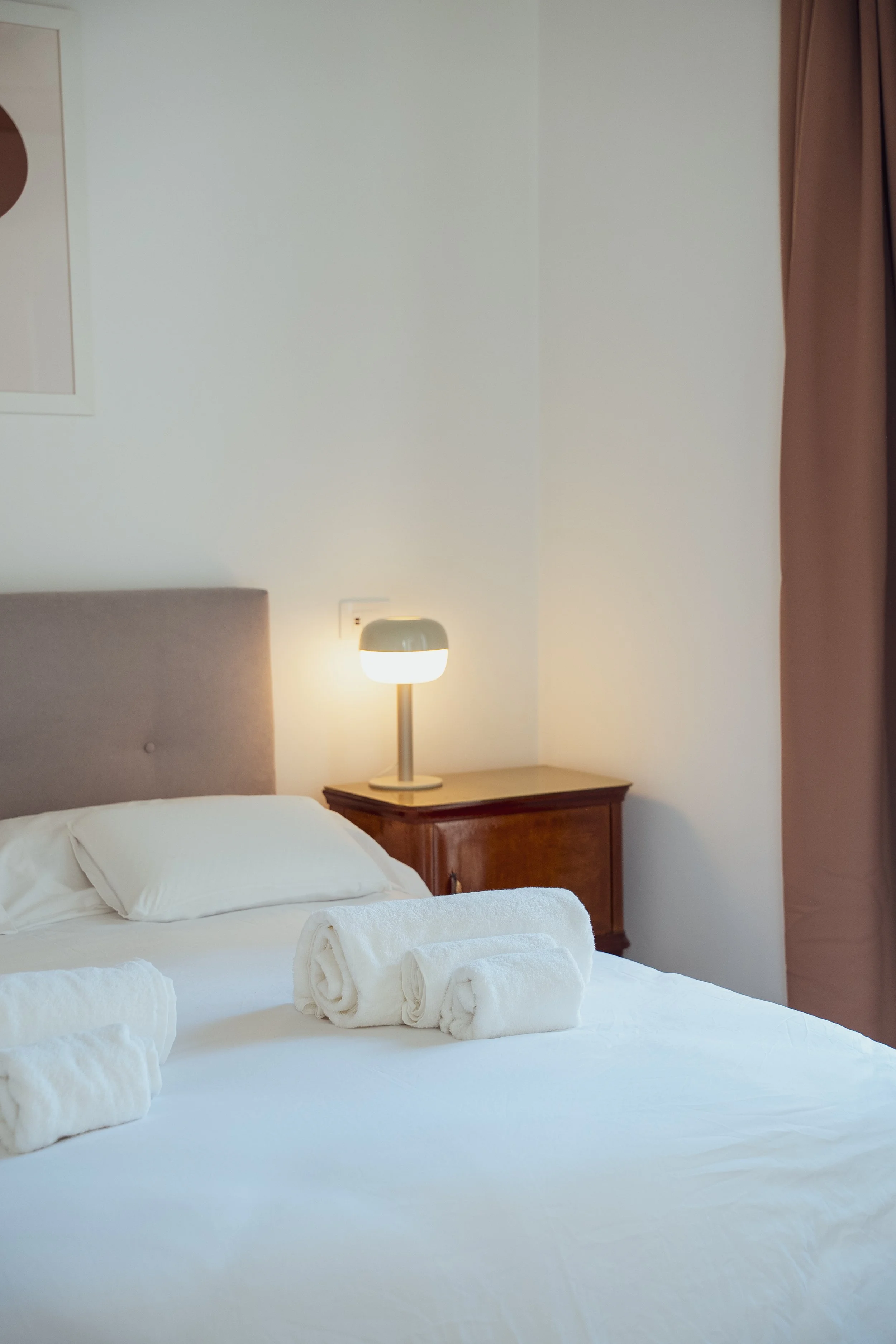 Camera da letto con letto matrimoniale con lenzuola bianche, cuscini e teli da rischio, lampada di tavolo su una nicchia di legno, pareti bianche e tendaggio marrone.