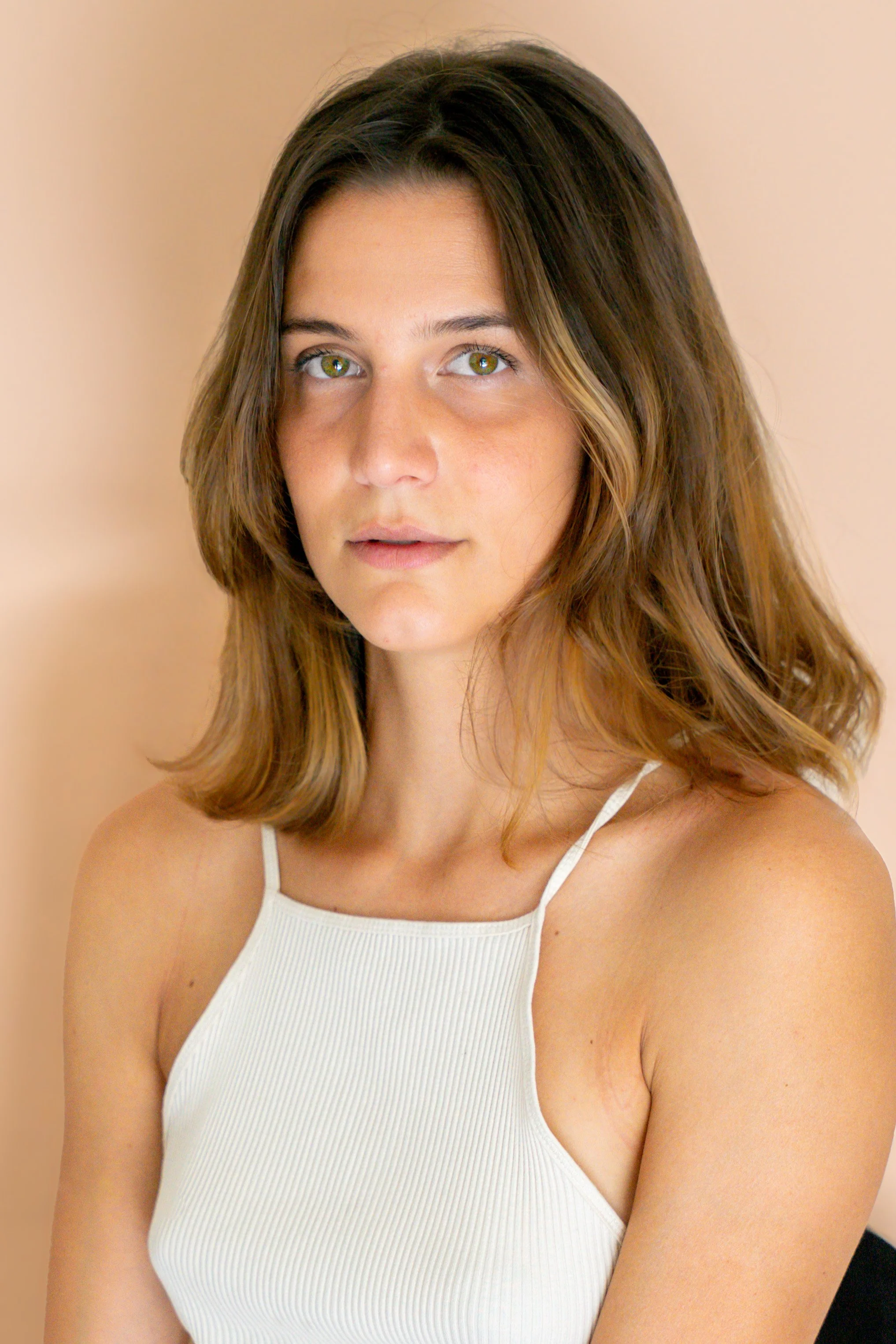 Ragazza con capelli castani, occhi verdi, camicia bianca a spallina sottile, sfondo beige.