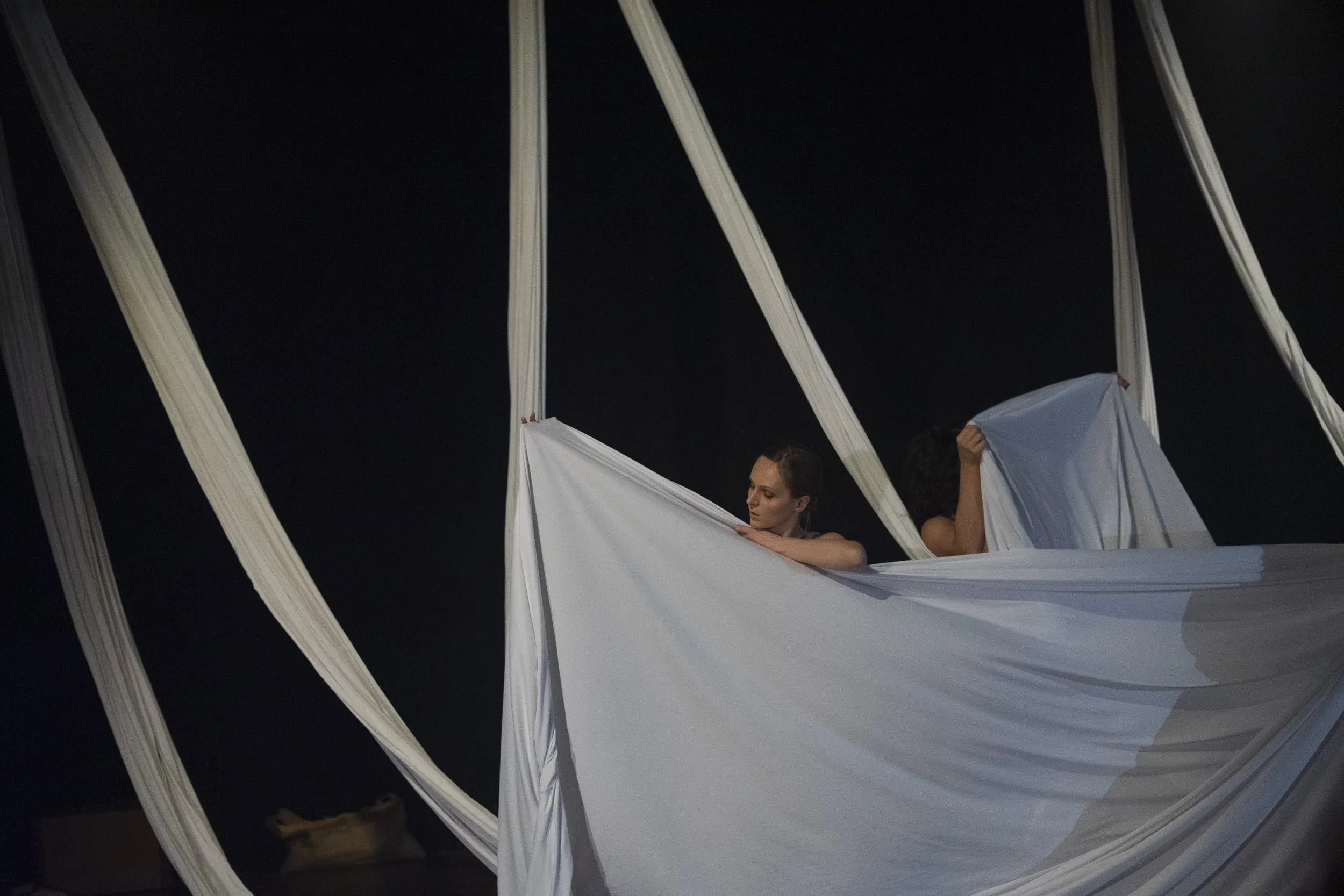 Due donne sospese in un'installazione artistica, circondate da lunghi veli bianchi contro uno sfondo nero.
