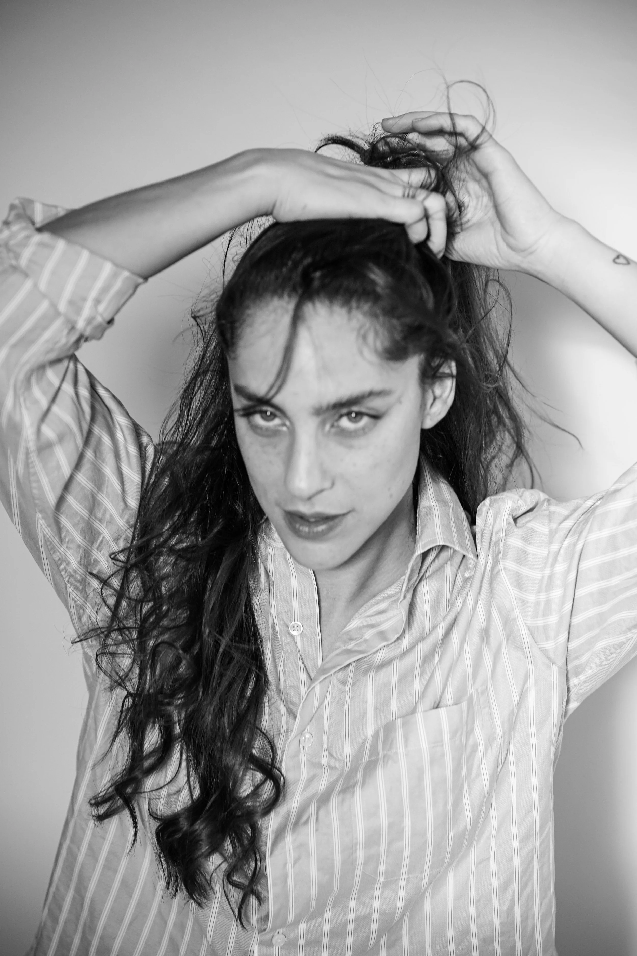 Ragazza con lunghi capelli ricci, che si sistema i capelli con le mani, indossa una camicia a righe e guarda verso l'obiettivo, in bianco e nero.