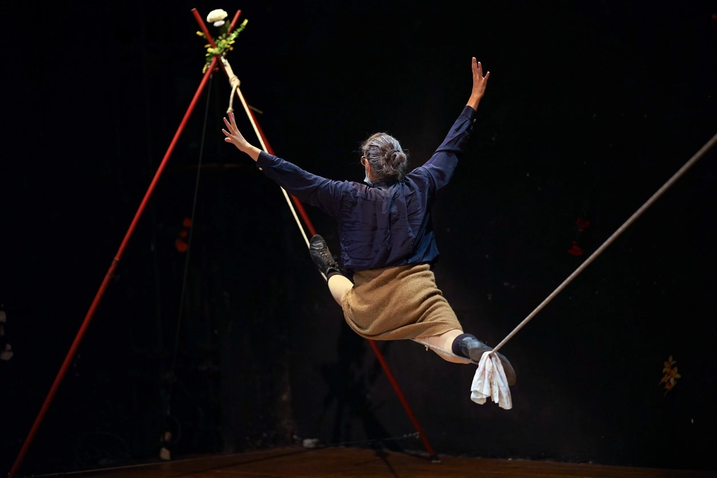 Una persona su una corda tesa, con i capelli grigi raccolti, che si esibisce in un numero di acrobazia o circo, con braccia alzate e sfondo nero.