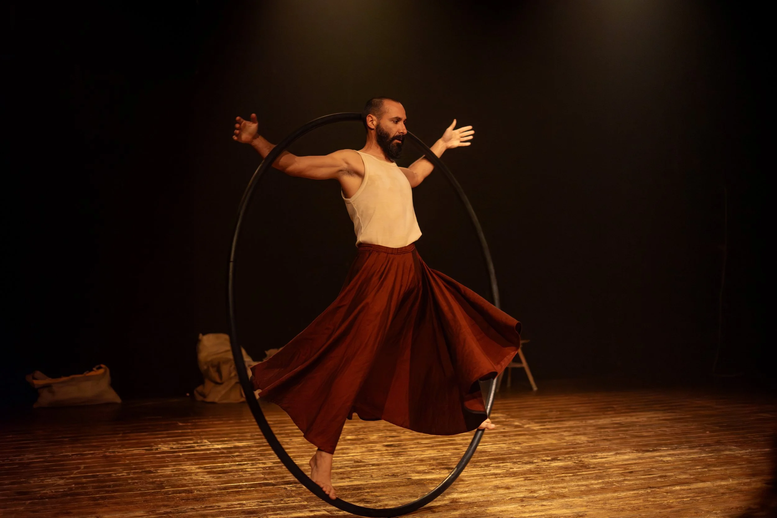 Un uomo con barba, vestito con pantaloni ampi color marrone e canotta beige, balla con un cerchio nero su un palco di legno, con un fascio di luce caldo.