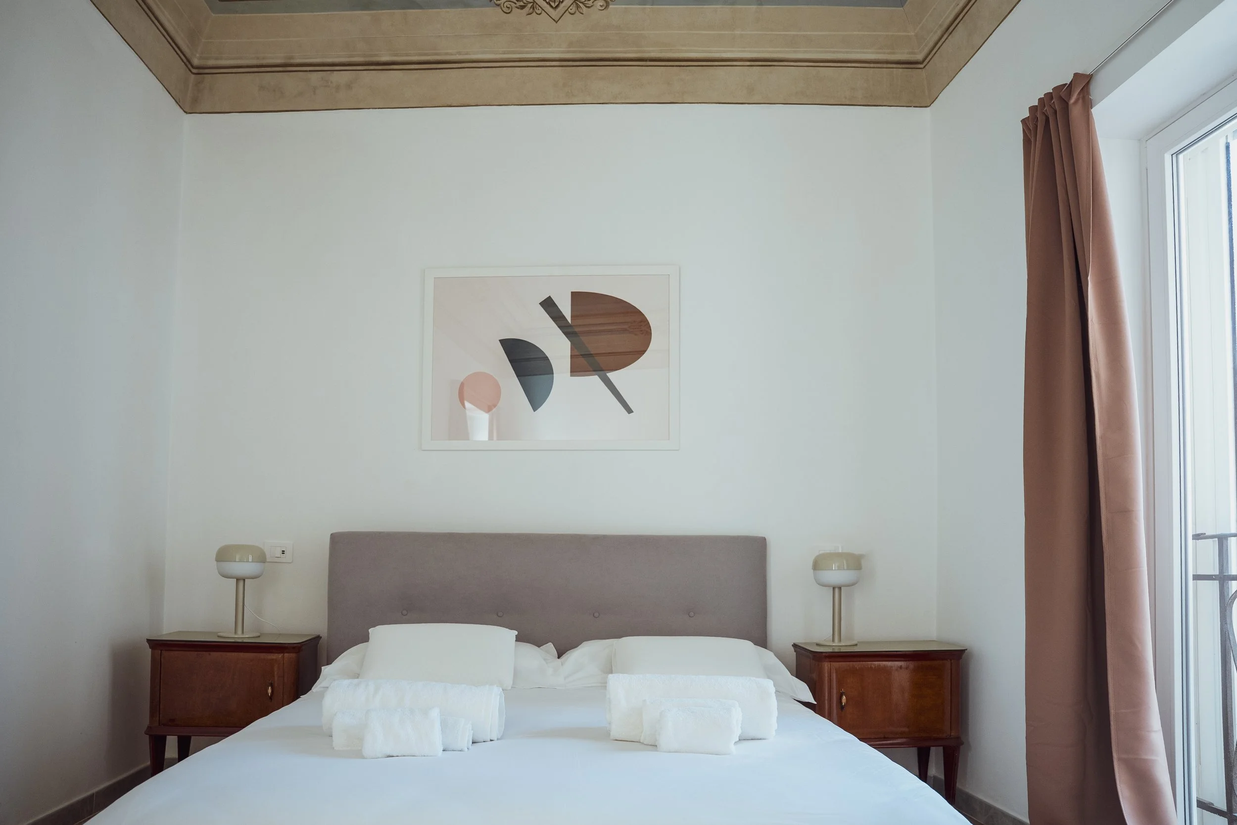 Camera da letto con letto matrimoniale, due comodini con lampade, quadro astratto sulla parete e tende color pelle accanto a una finestra