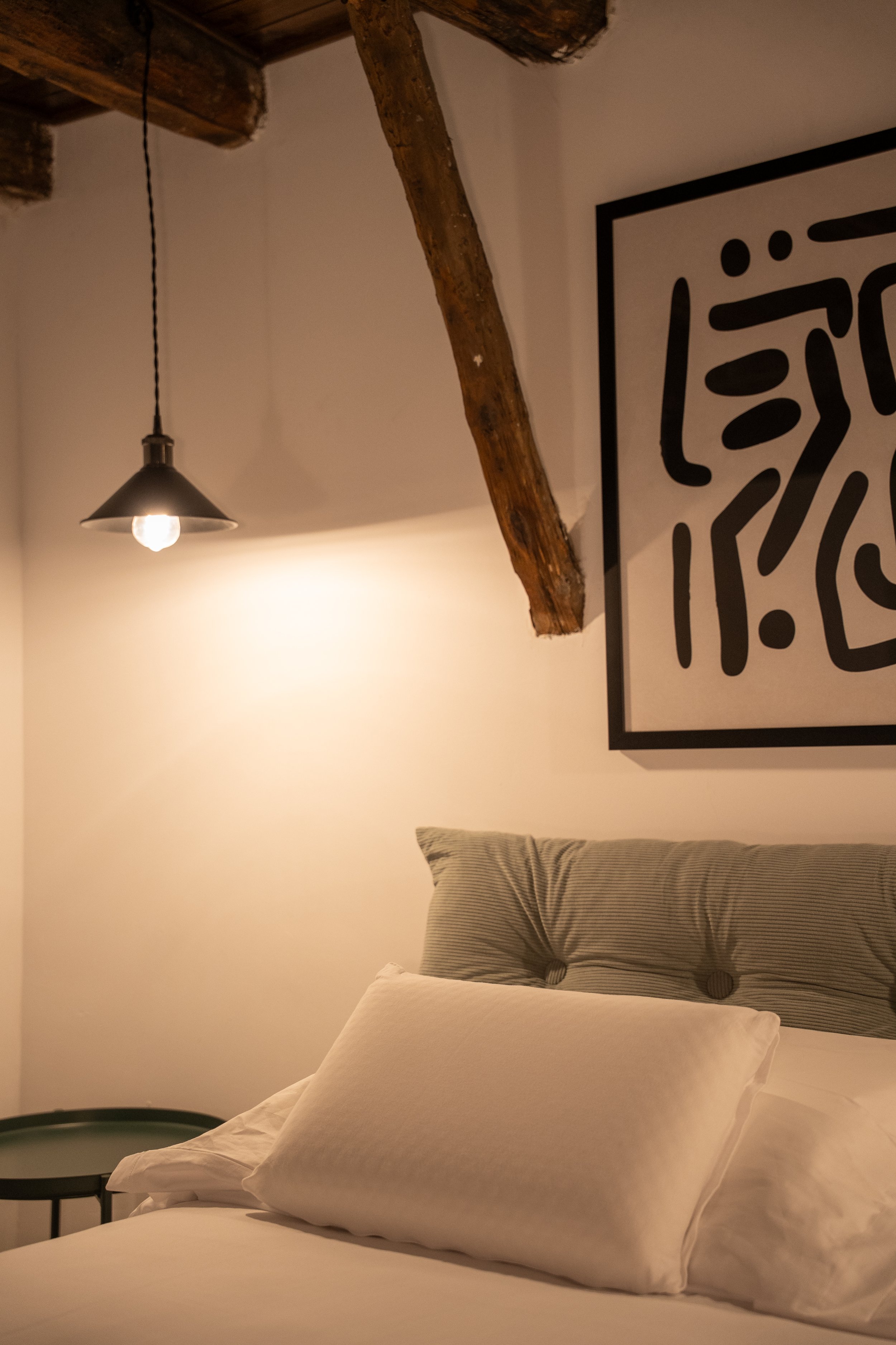 Letto matrimoniale con cuscino bianco e testiera imbottita grigia, lampada appesa con luce calda, parete con quadro astratto nero e bianco, soffitto con travi di legno.