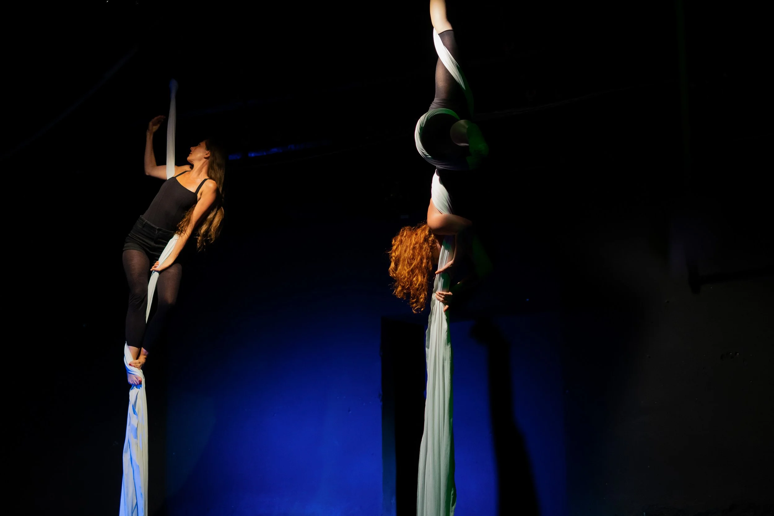 Due donne si esibiscono in una performance di acrobatica aerea, appese a lunghe stoffe color avorio in un ambiente buio con illuminazione soffusa.