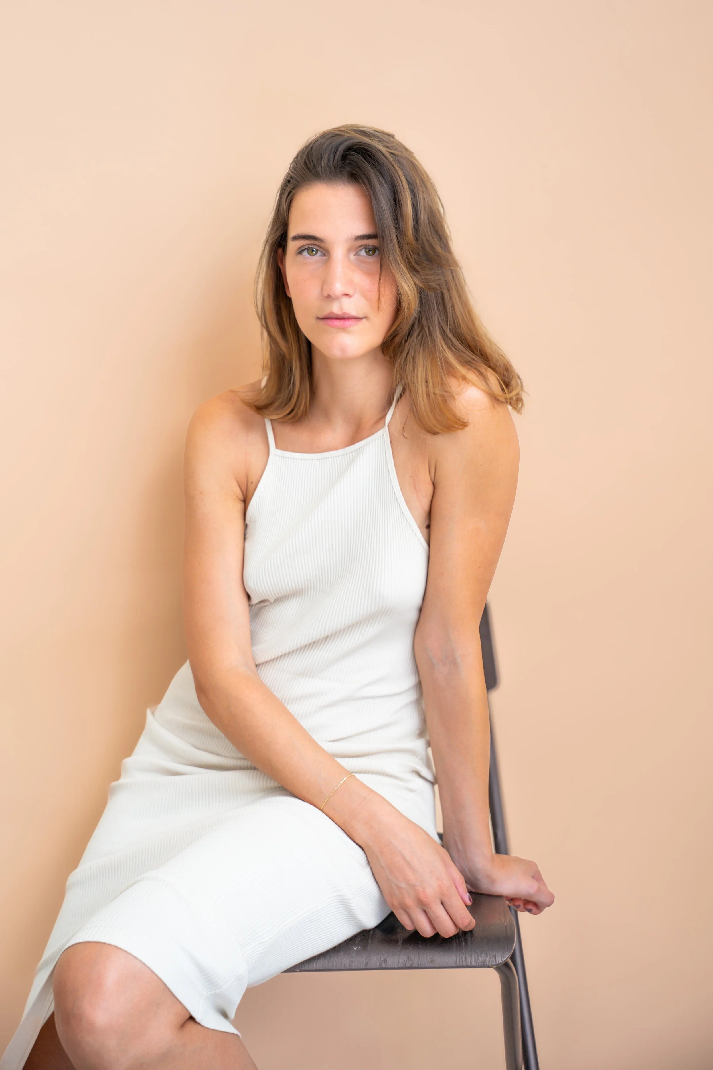 Una giovane donna seduta su una sedia, con capelli castani mossi, indossa un vestito bianco senza spalline, posa di fronte a uno sfondo beige.