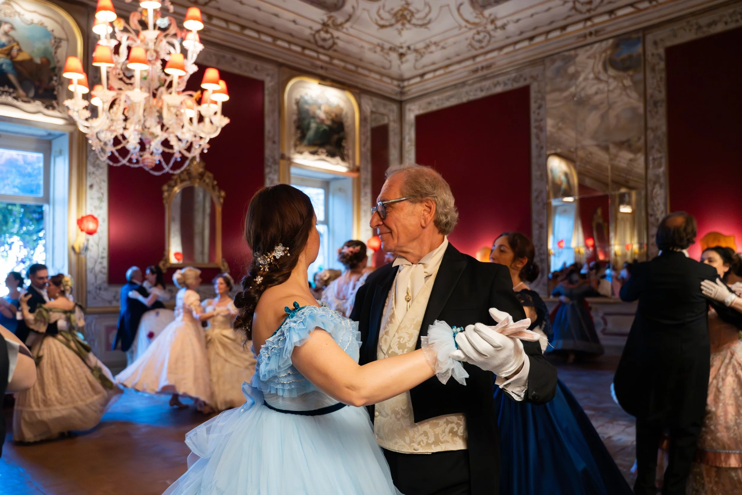 Un uomo anziano e una donna danzano insieme in un salone elegante decorato con specchi dorati, dipinti e un lampadario di cristallo con lampade rosse, in presenza di altri coppie che ballano.