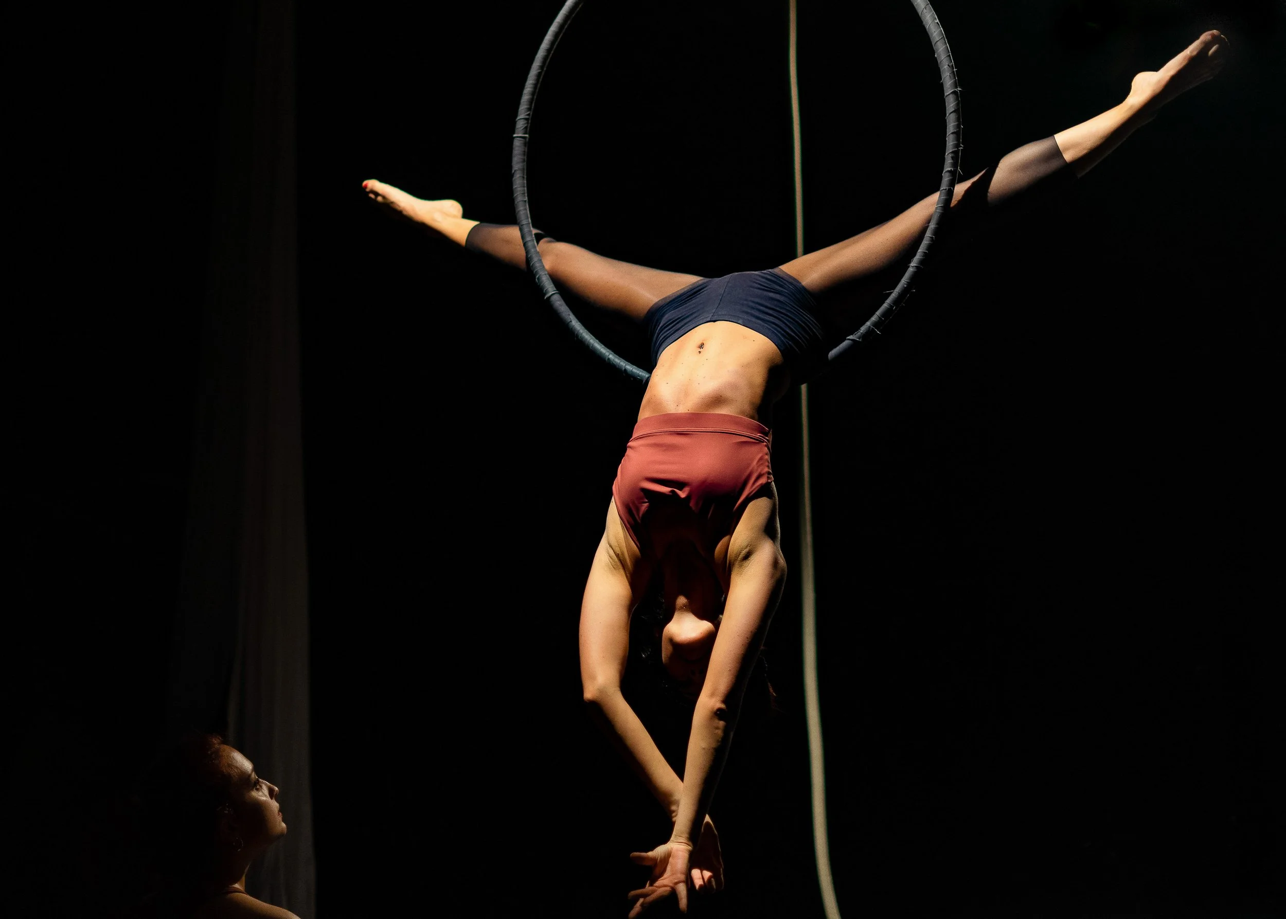 Una donna sta esibendo un acrobazia con un cerchio aereo sul palco di un circo o teatro, con un'altra donna che la guarda.