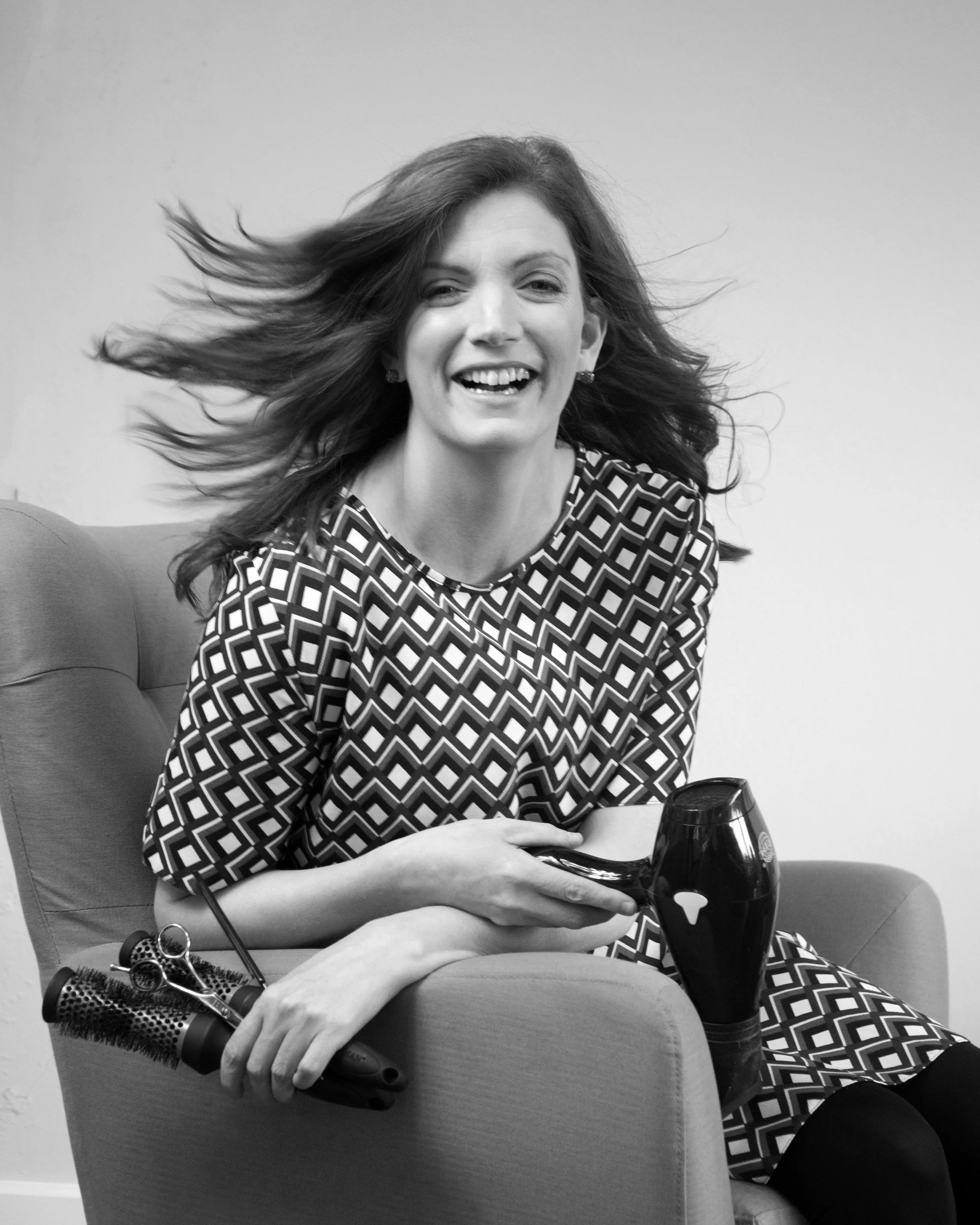 A woman with long hair smiling and holding a wine glass, sitting on a chair. Hair is blowing to the side, and she is wearing a patterned dress.