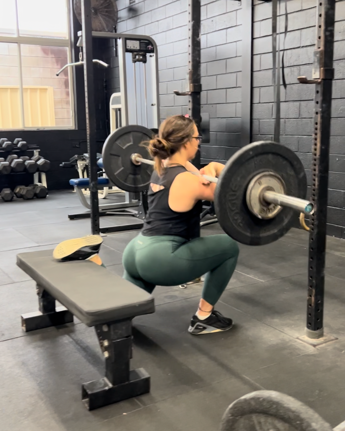 Woman performing a Bulgarian split squat with a barbell in a gym, positioned on a bench with black weights on a black squat rack.