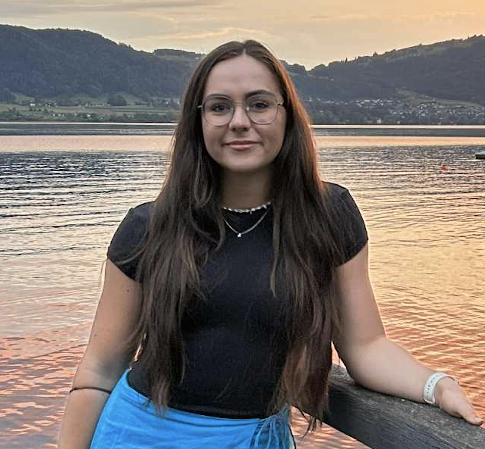 A young woman with long brown hair and glasses standing by a lake at sunset, wearing a black t-shirt, blue skirt, and necklaces.