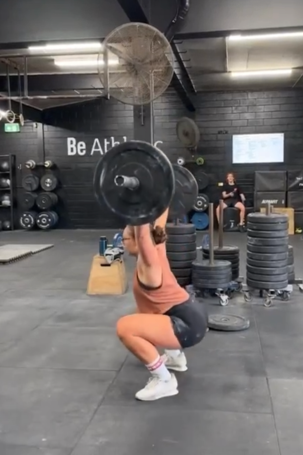 A woman is performing a back squat exercise with a barbell in a gym. She is in a squat position with her knees bent and hips lowered, holding the barbell overhead. The gym has stacked weights, exercise equipment, and an individual sitting on a bench in the background.