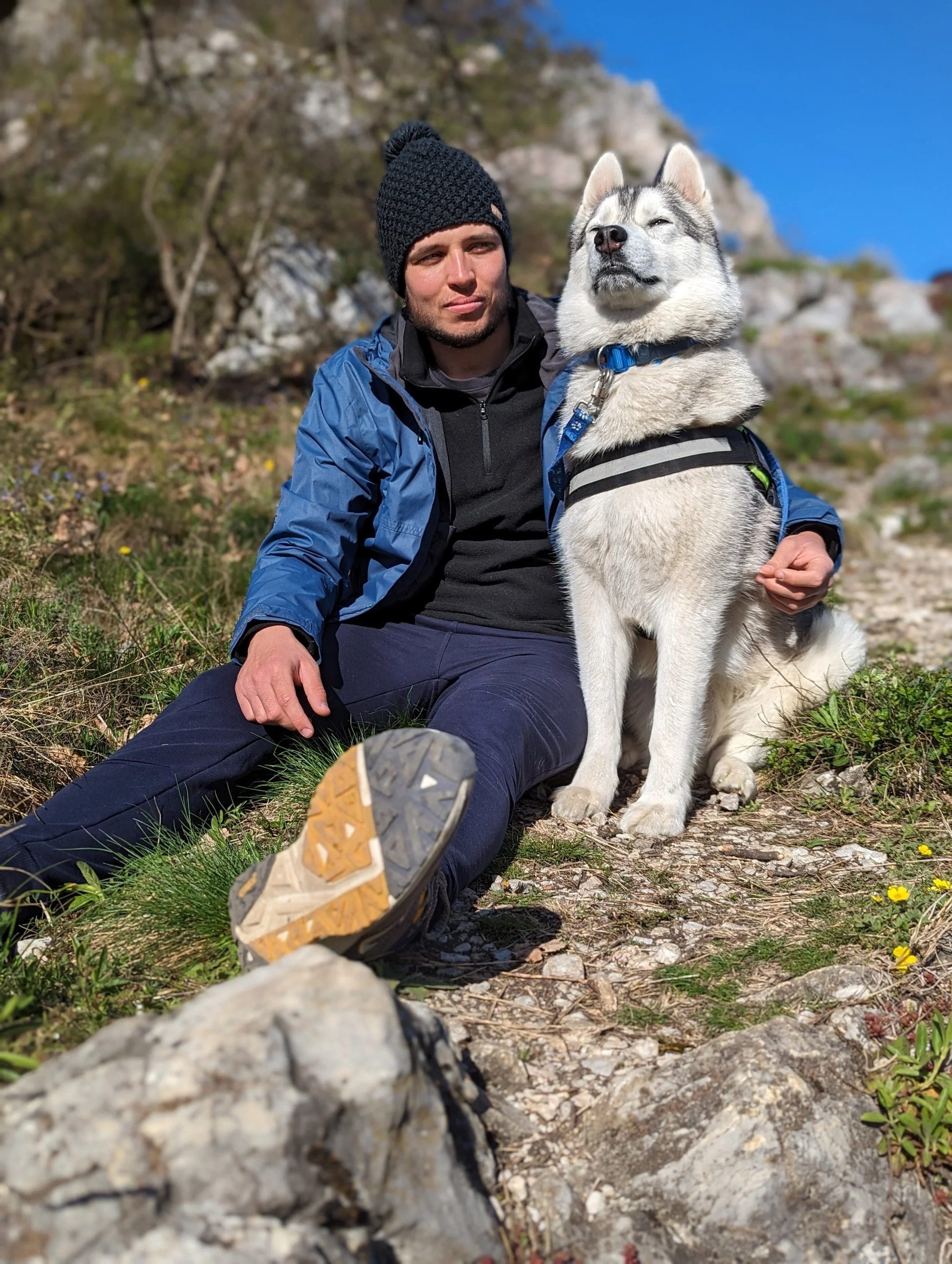 A man in outdoor hiking gear sitting on a rocky trail next to a Siberian Husky dog during a hike in a mountainous area with green vegetation and a clear blue sky.