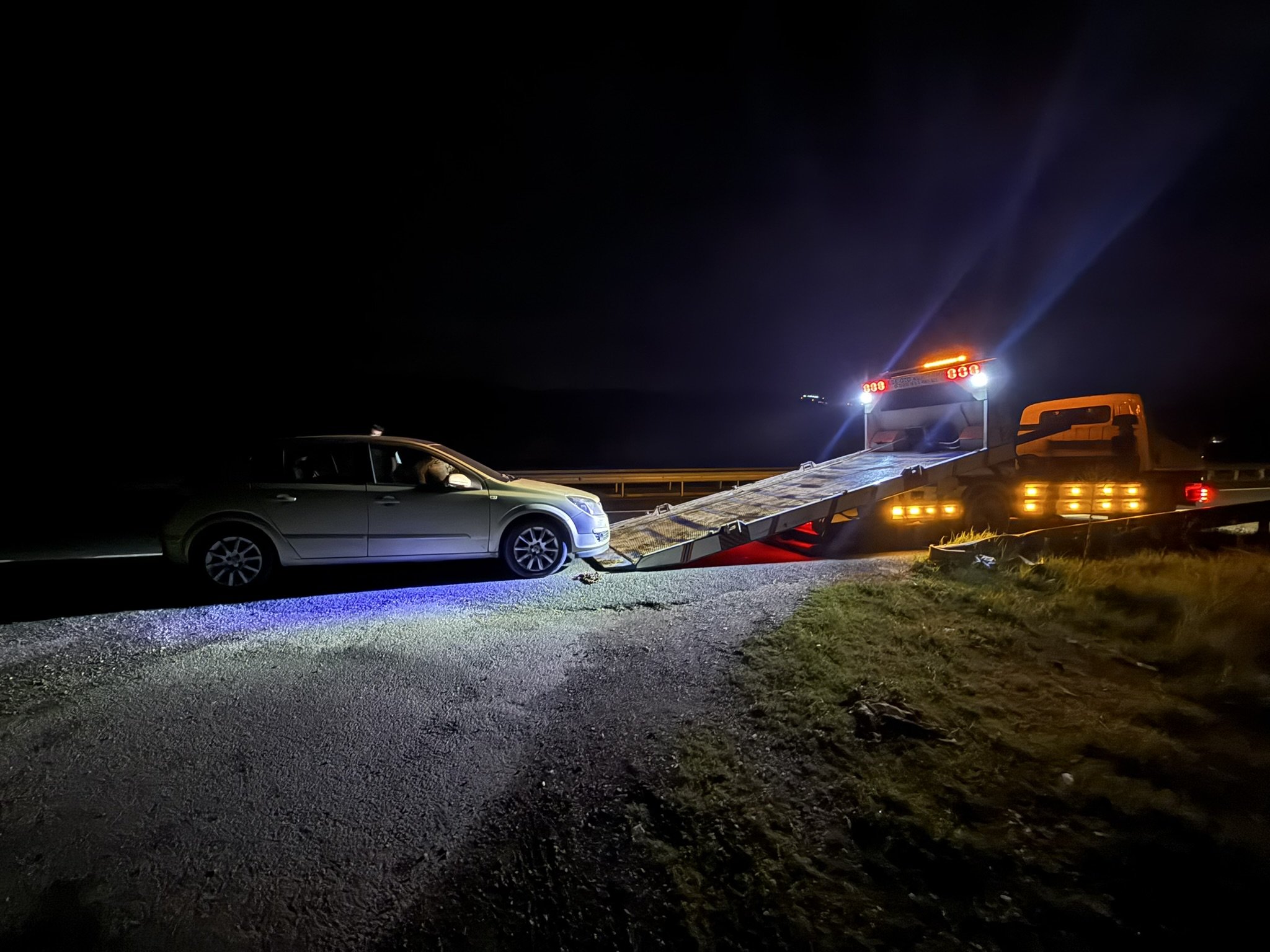 Gece vakti, karanlık bir yol kenarında, ışıkları yanıp sönen bir çekici kamyon gümüş renkli bir station wagon'u çekiyor.