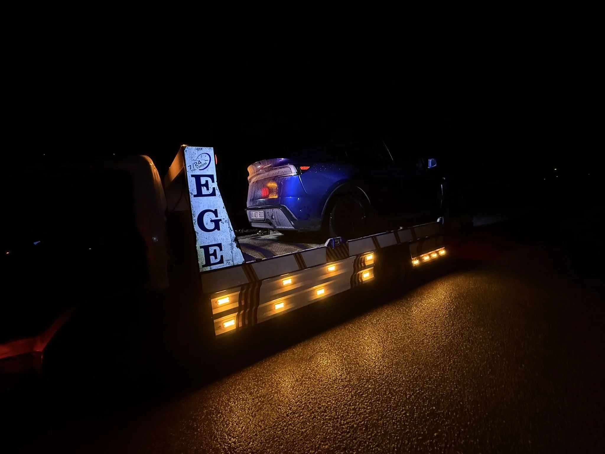 Gece vakti, mavi bir araba taşıyan bir çekici kamyonu. Kamyonun yan tarafında, aracın farlarıyla aydınlatılmış, EGE harflerini gösteren yansıtıcı bir levha bulunuyor.