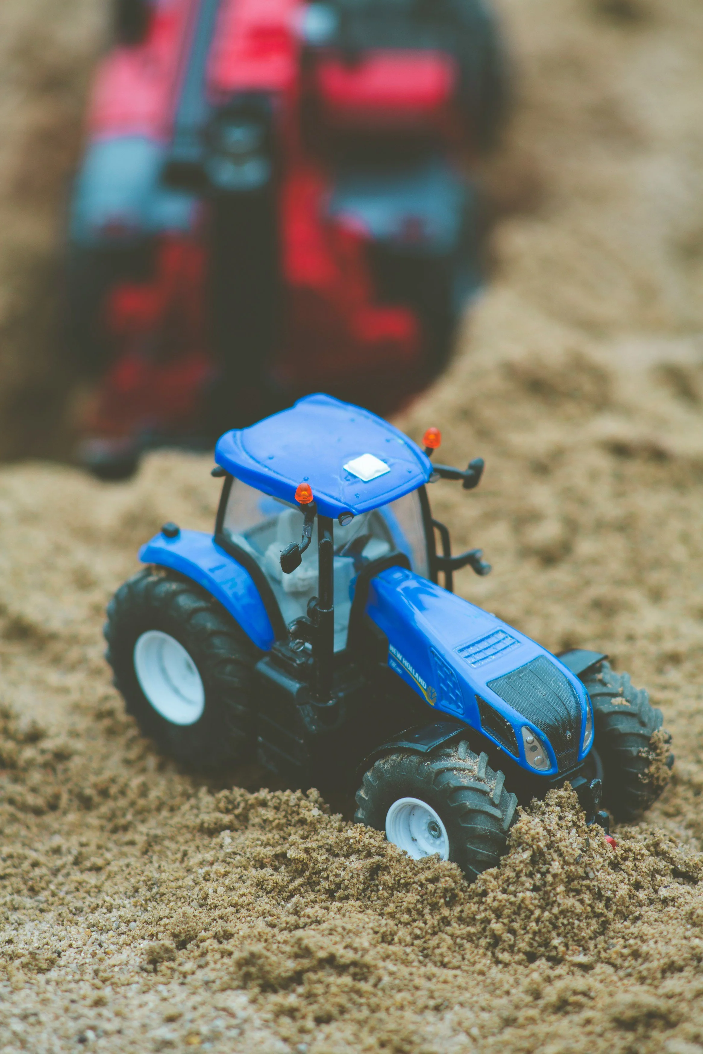Close-up of a blue toy tractor on sandy ground, with a blurred red toy vehicle in the background.