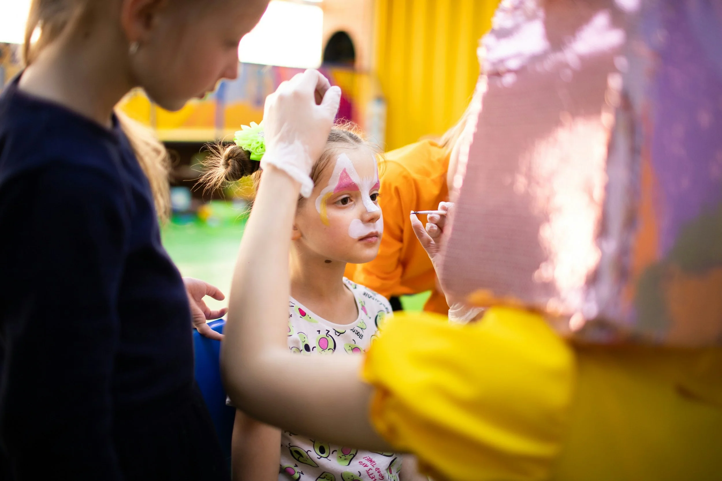 A young girl with her face painted like a rainbow, getting her face painted by an artist in an outdoor setting.