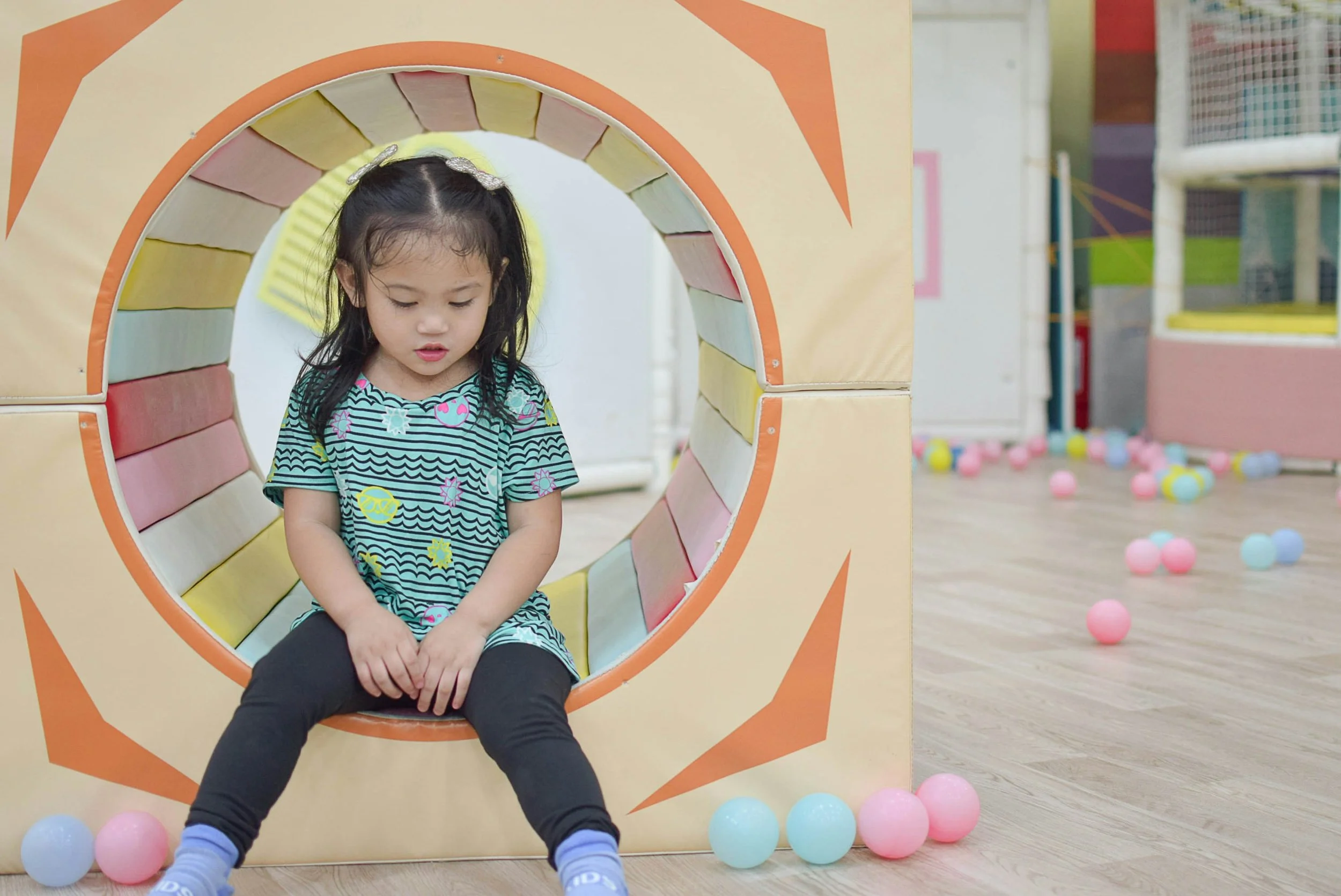 A young girl sitting inside a colorful play tunnel with pastel balls scattered around on a wooden floor, in an indoor play area.