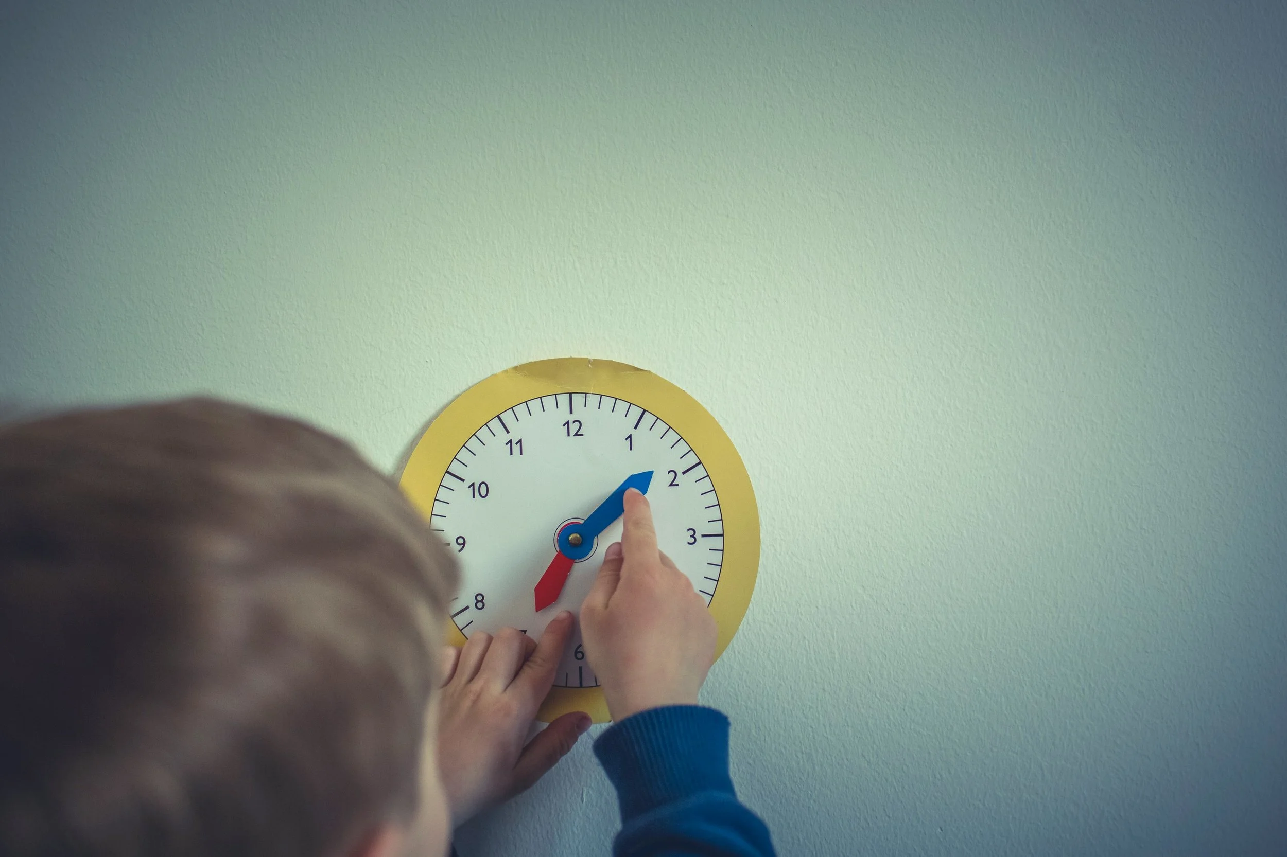 A young child with light-colored hair adjusting a round open-face clock on a white wall. The clock has a yellow frame, blue hour hand, red minute hand, and standard black numbers.