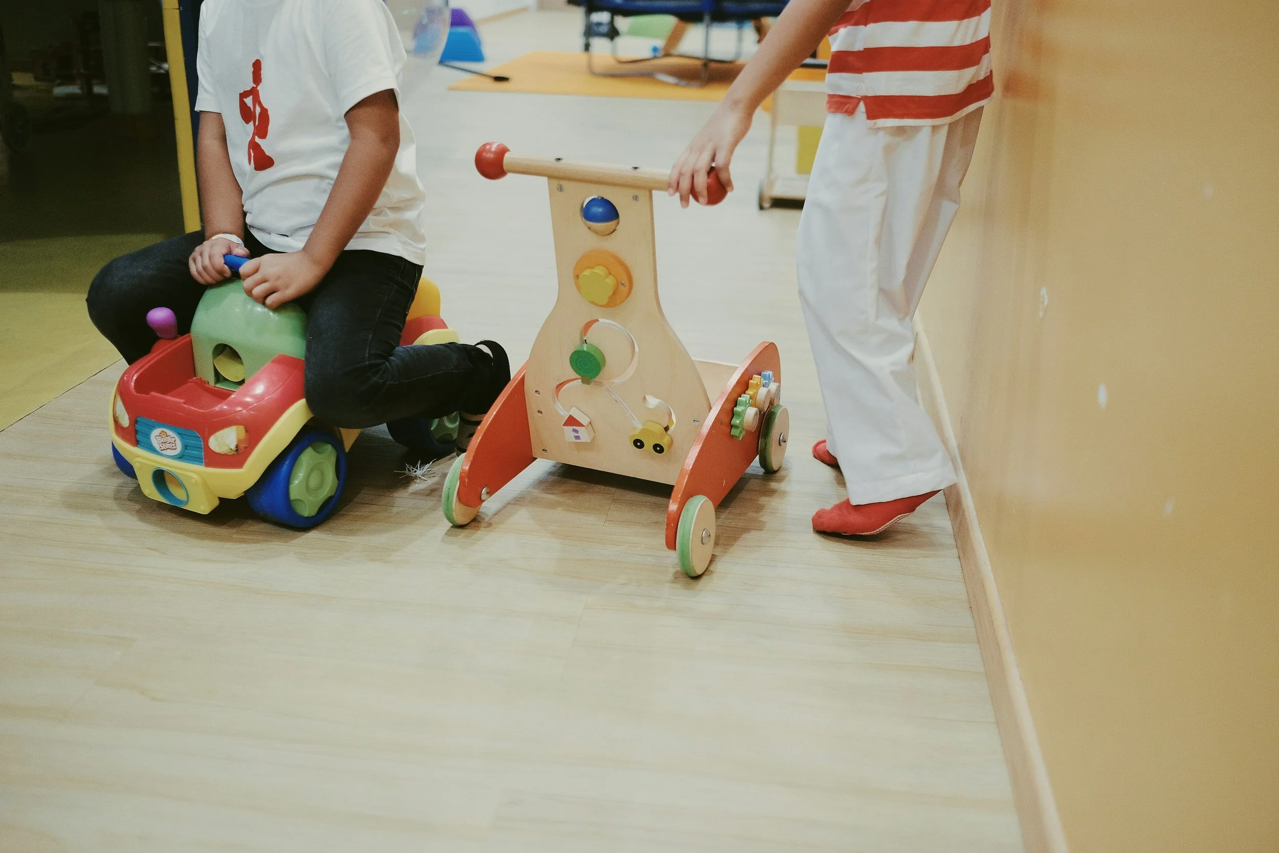 Two children playing with toys indoors. One child is sitting on a colorful ride-on toy, and the other is pushing a wooden toy with wheels and various attached elements.
