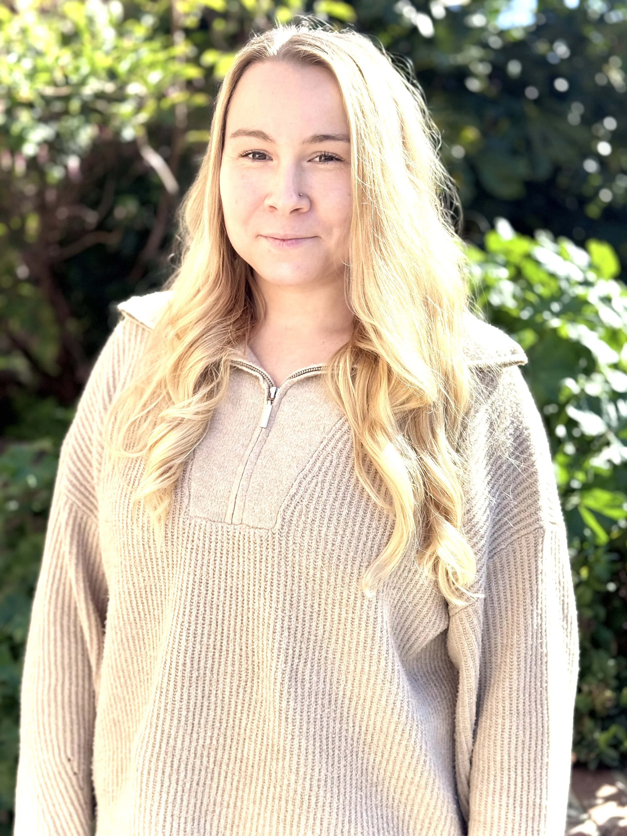 A young woman with long, wavy blonde hair standing outdoors in front of green foliage, wearing a beige quarter-zip sweater.