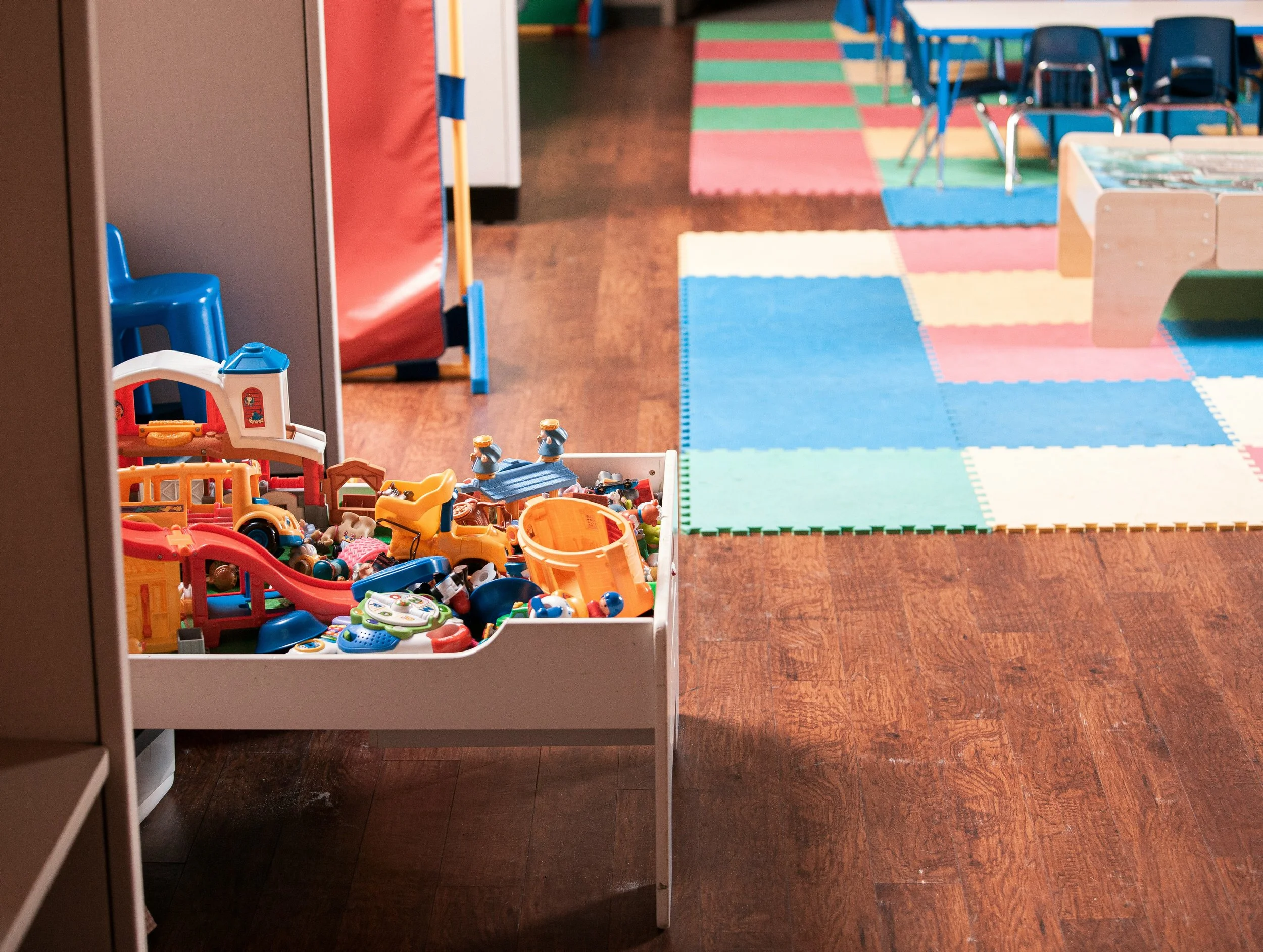 A toy bin filled with small plastic toys and figurines in a playroom with a colorful foam mat and small chairs and tables.