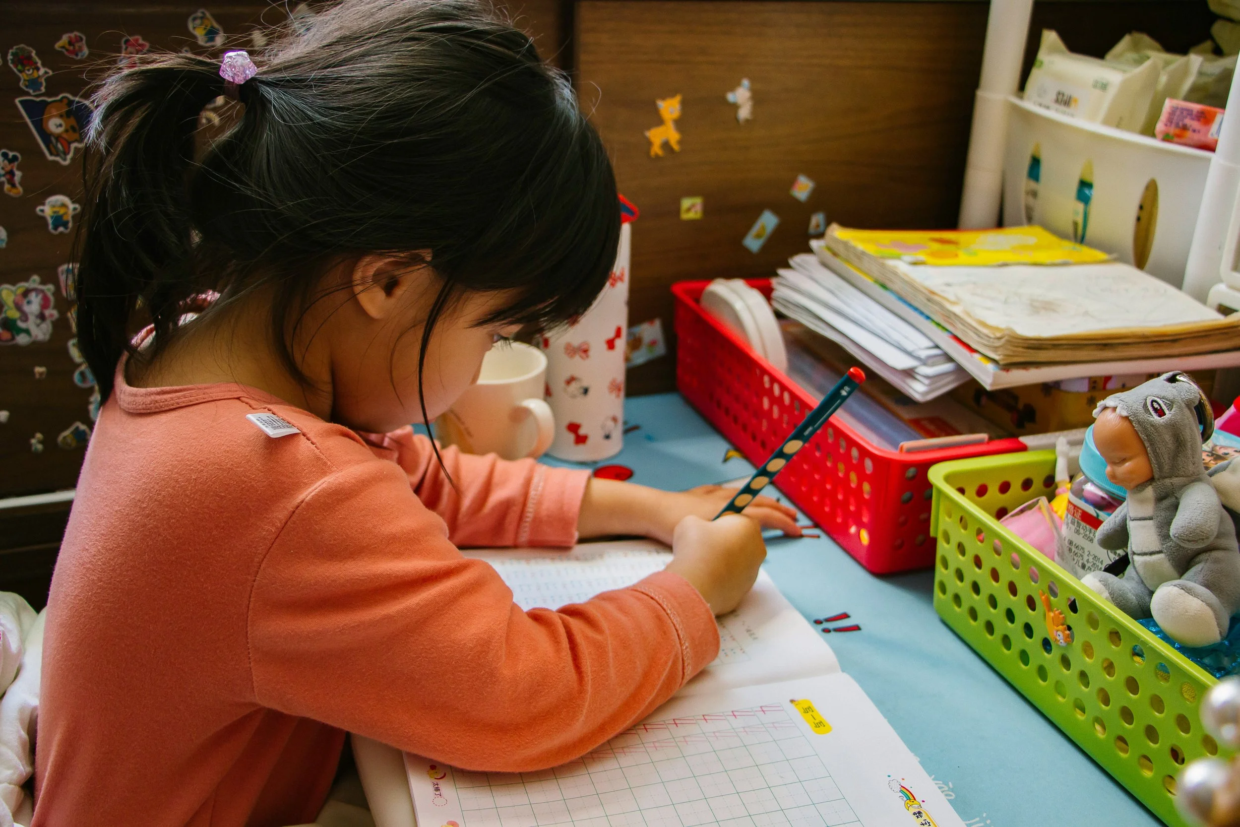 A young girl with dark hair in a ponytail, wearing a peach-colored shirt, sitting at a desk and doing homework with a pencil. The desk has colorful storage containers, papers, and a plush toy dressed in a gray hoodie with a face mask nearby.