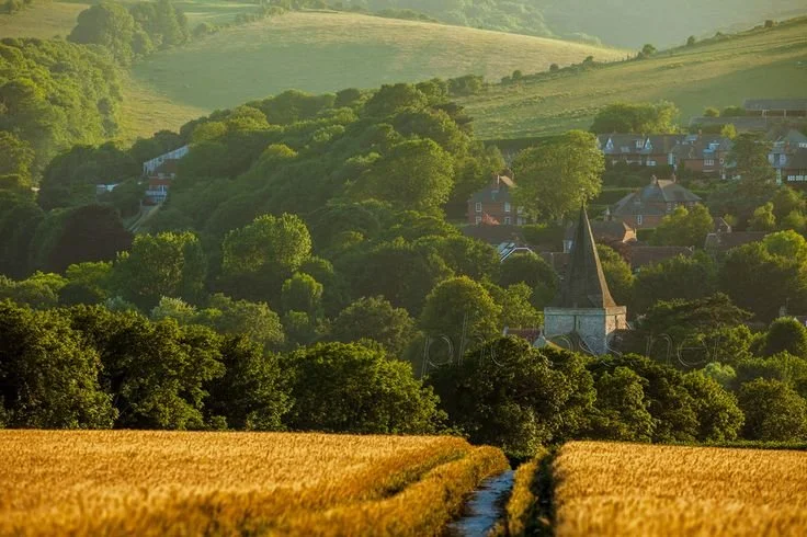 A lush green rural landscape with a village, a church with a tall steeple, surrounded by rolling hills and fields of crops.
