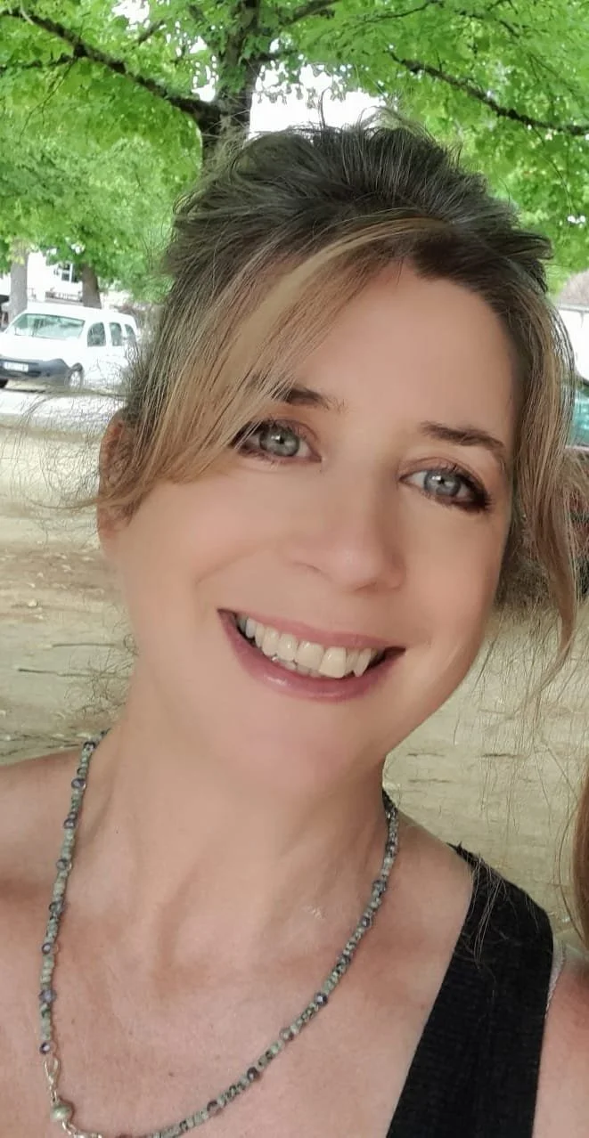 Close-up of a smiling woman with blonde hair, blue eyes, and light skin, standing outdoors under a green leafy tree with cars and a sidewalk in the background.