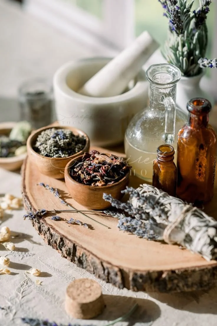 A wooden tray with small bowls of dried herbs, a bundle of dried lavender, a glass jar of liquid, and two small brown bottles, surrounded by dried flowers and herbs.