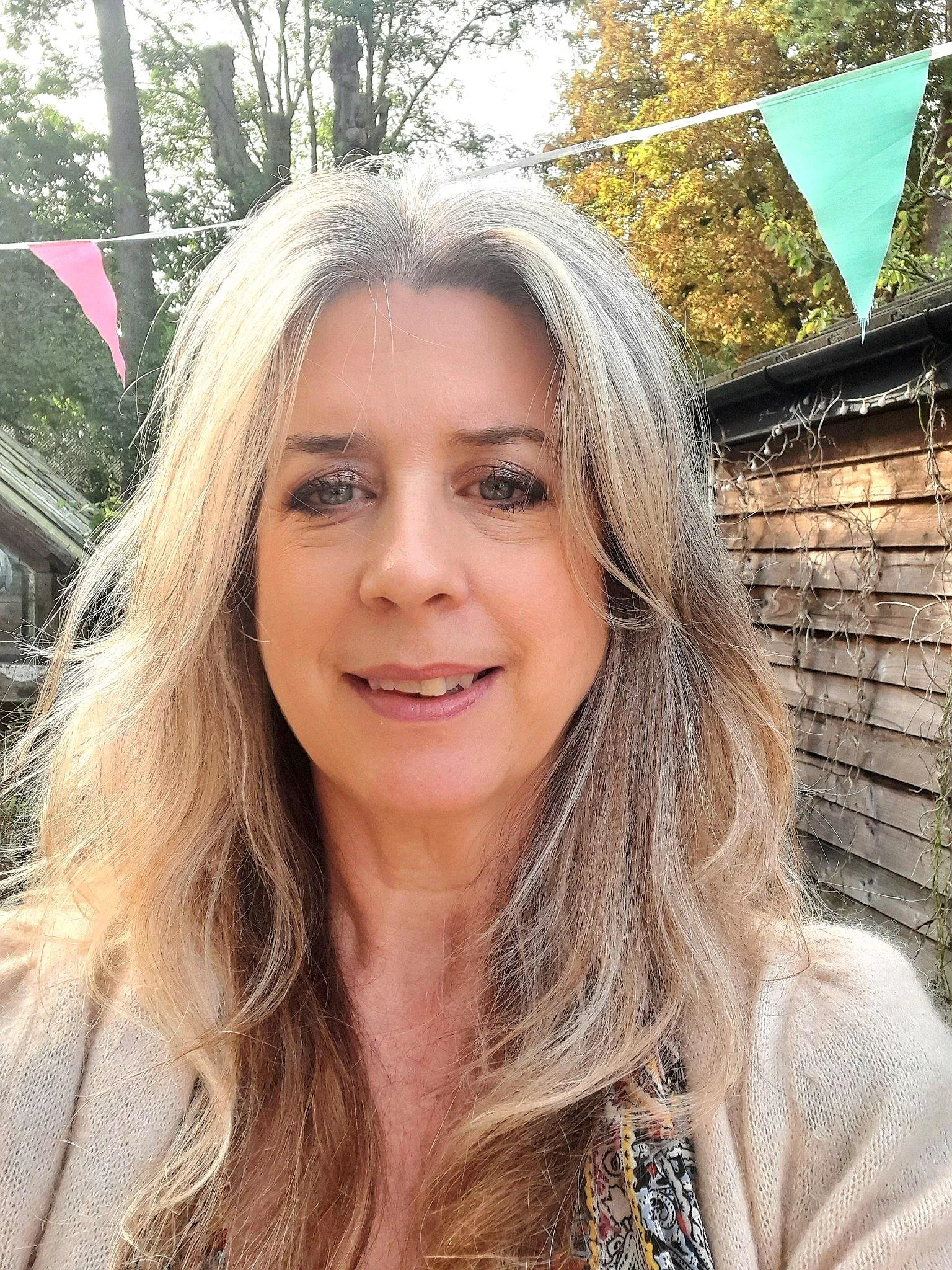 A smiling woman with long, wavy blonde hair takes a selfie outdoors with trees and colorful bunting in the background.