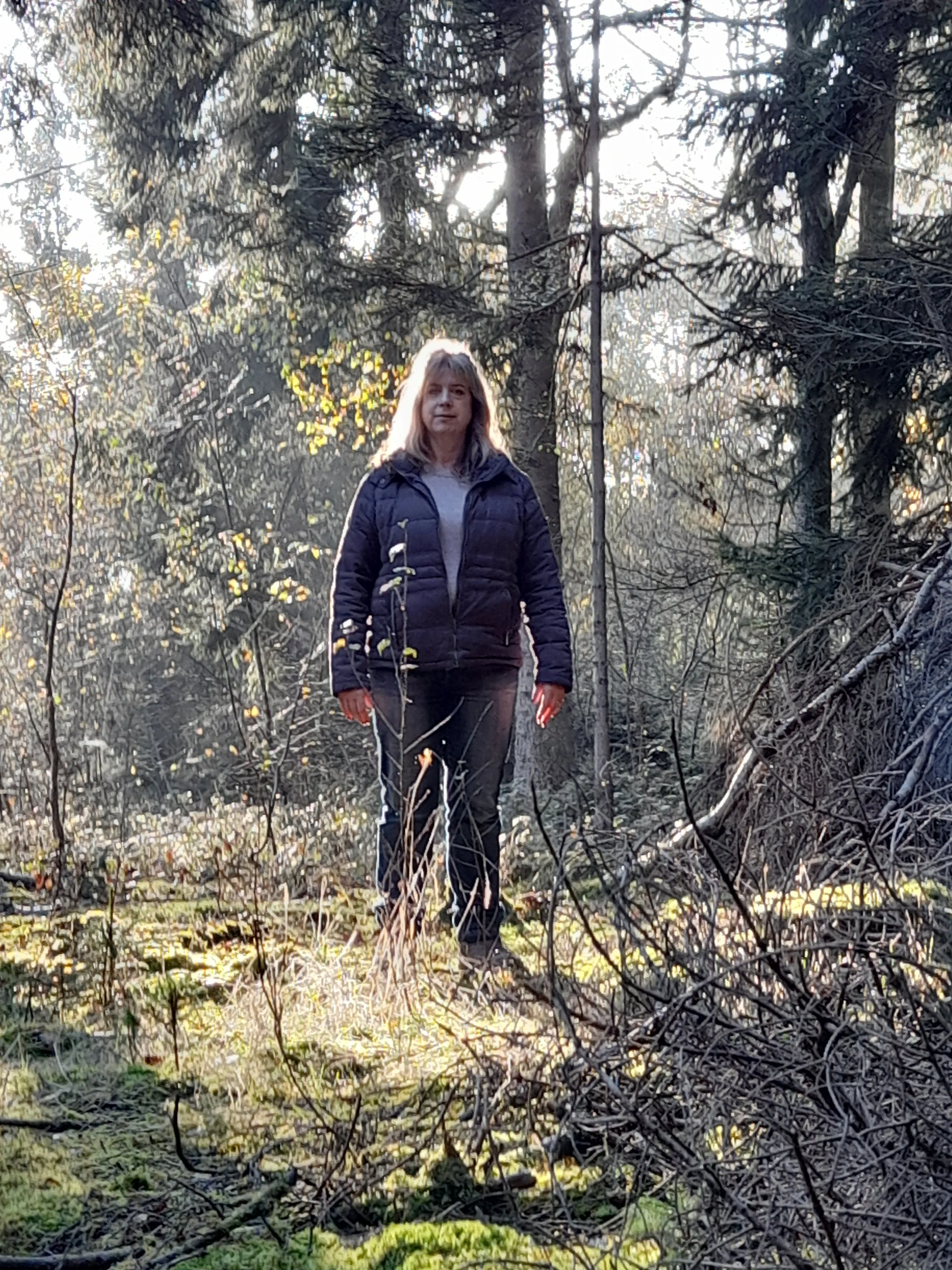 A woman standing outdoors in a forested area with sunlight filtering through the trees.