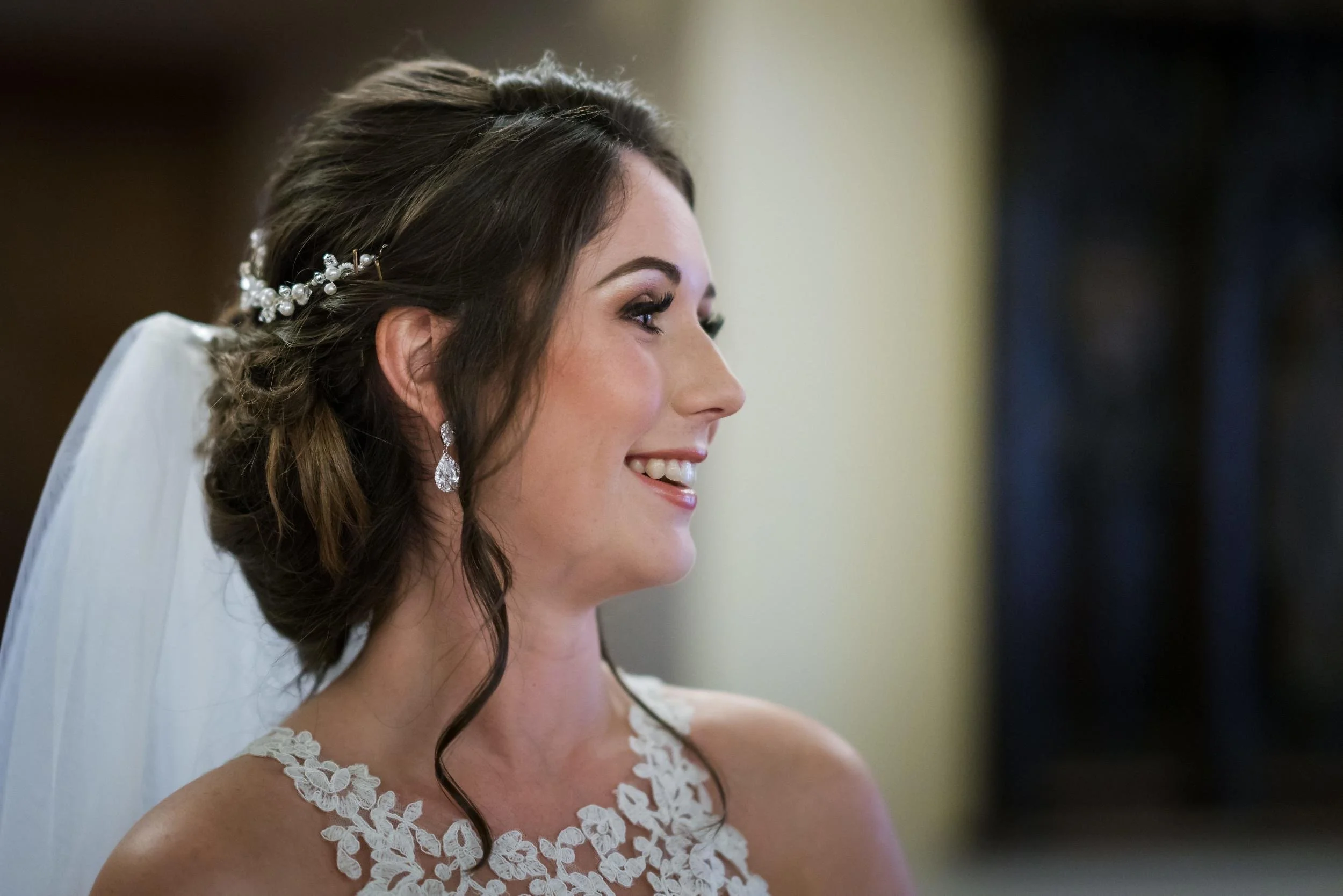 Side profile of a smiling bride with dark hair styled in waves, wearing a lace wedding dress, pearl hair accessory, and dangling diamond earrings.