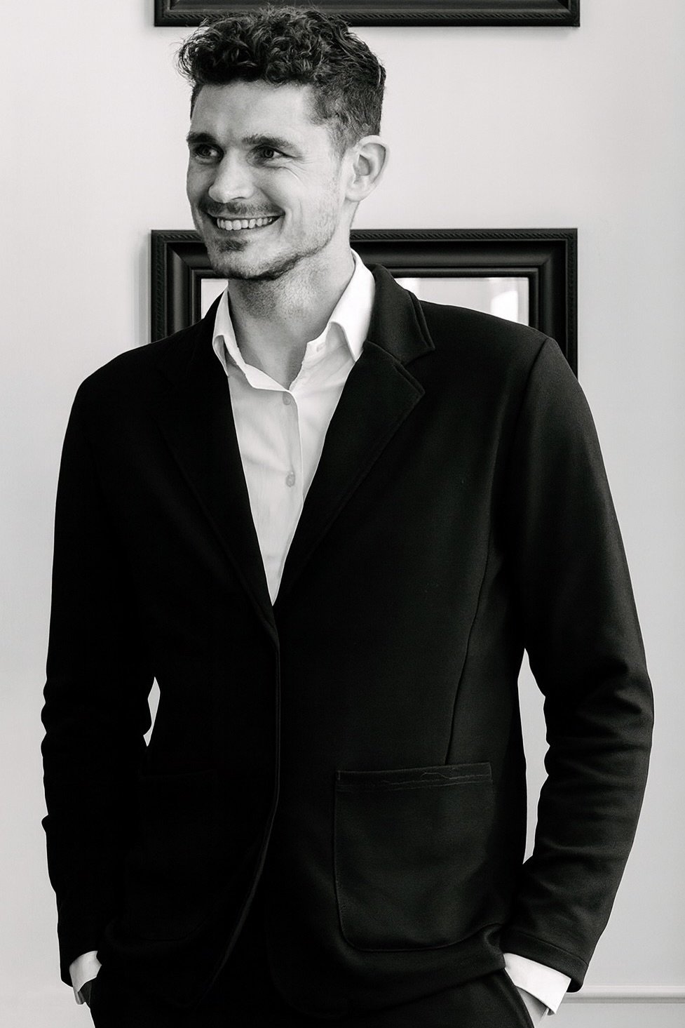 A smiling man in a black blazer and white shirt standing in front of framed pictures on a white wall.